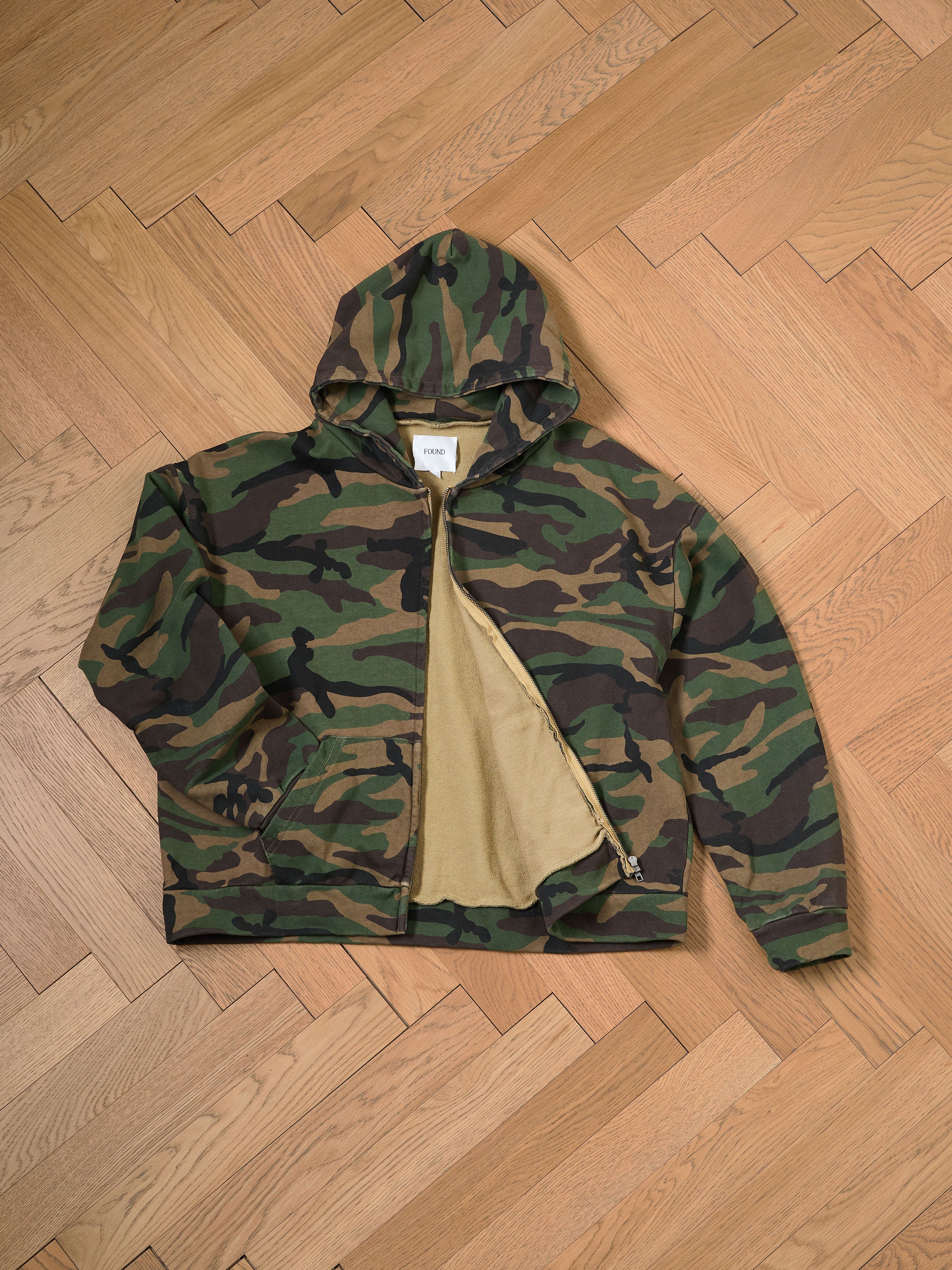 The Washed Camouflage Zip Hoodie by FOUND, featuring a hood and crafted from soft French terry cotton, lies open on a wooden herringbone floor.