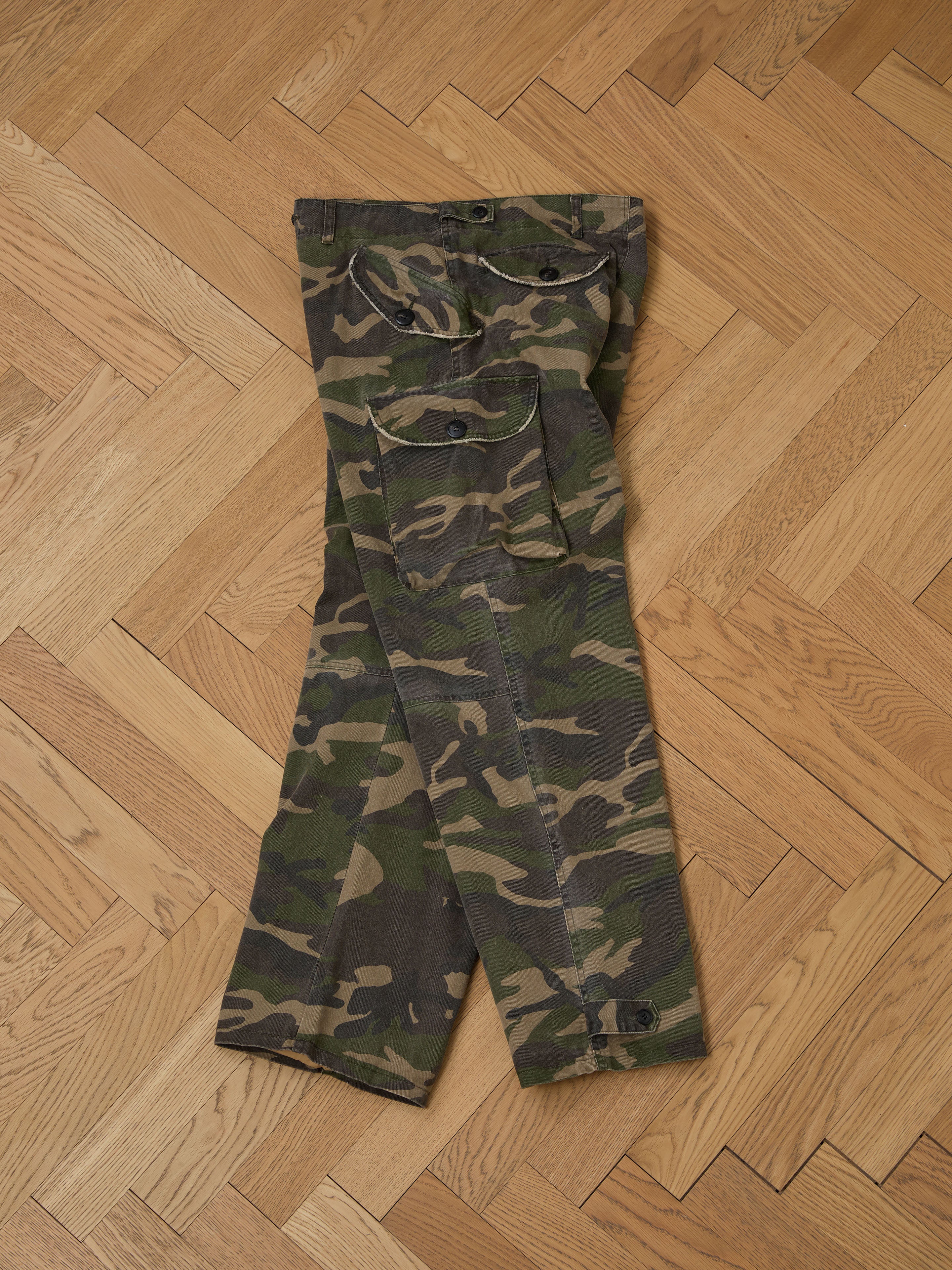 A pair of FOUND Camouflage Twill Utility Cargo Pants with a vintage look lies neatly folded on the wooden herringbone-patterned floor.