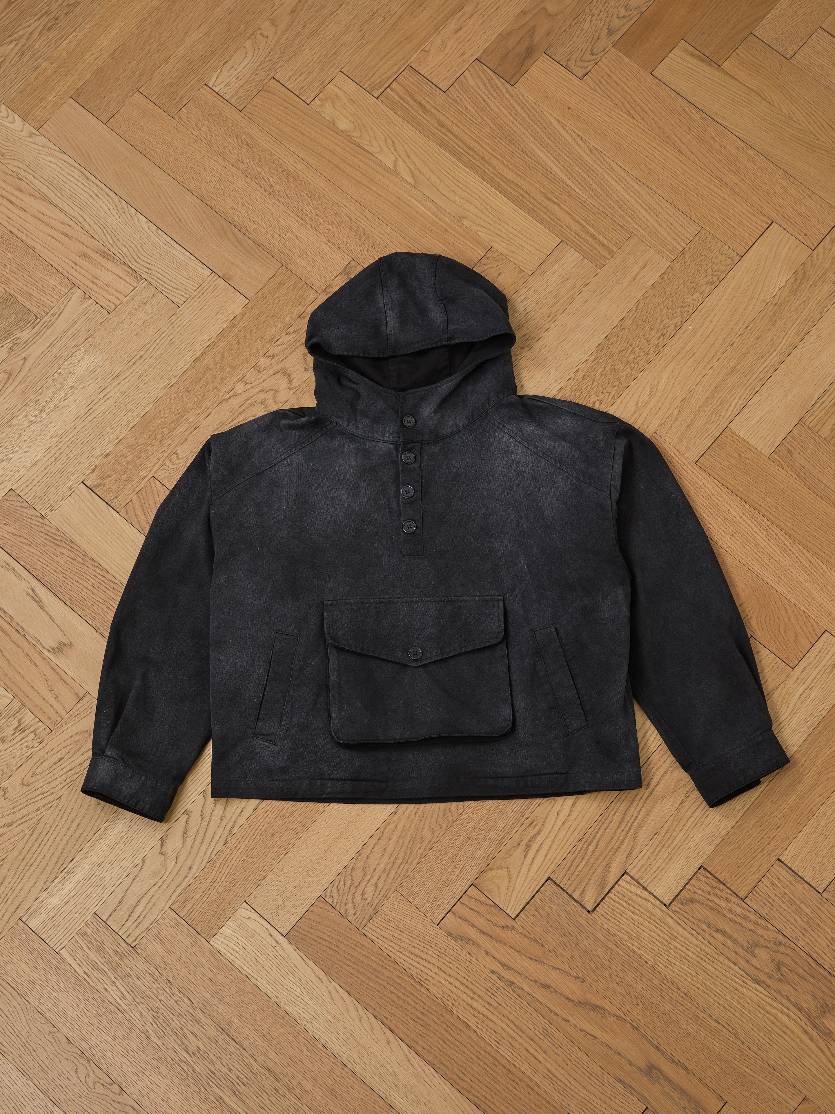 The Washed Twill Pocket Anorak Jacket by FOUND is a vintage black piece featuring a buttoned front and large cotton twill pocket, displayed on a wooden herringbone floor.