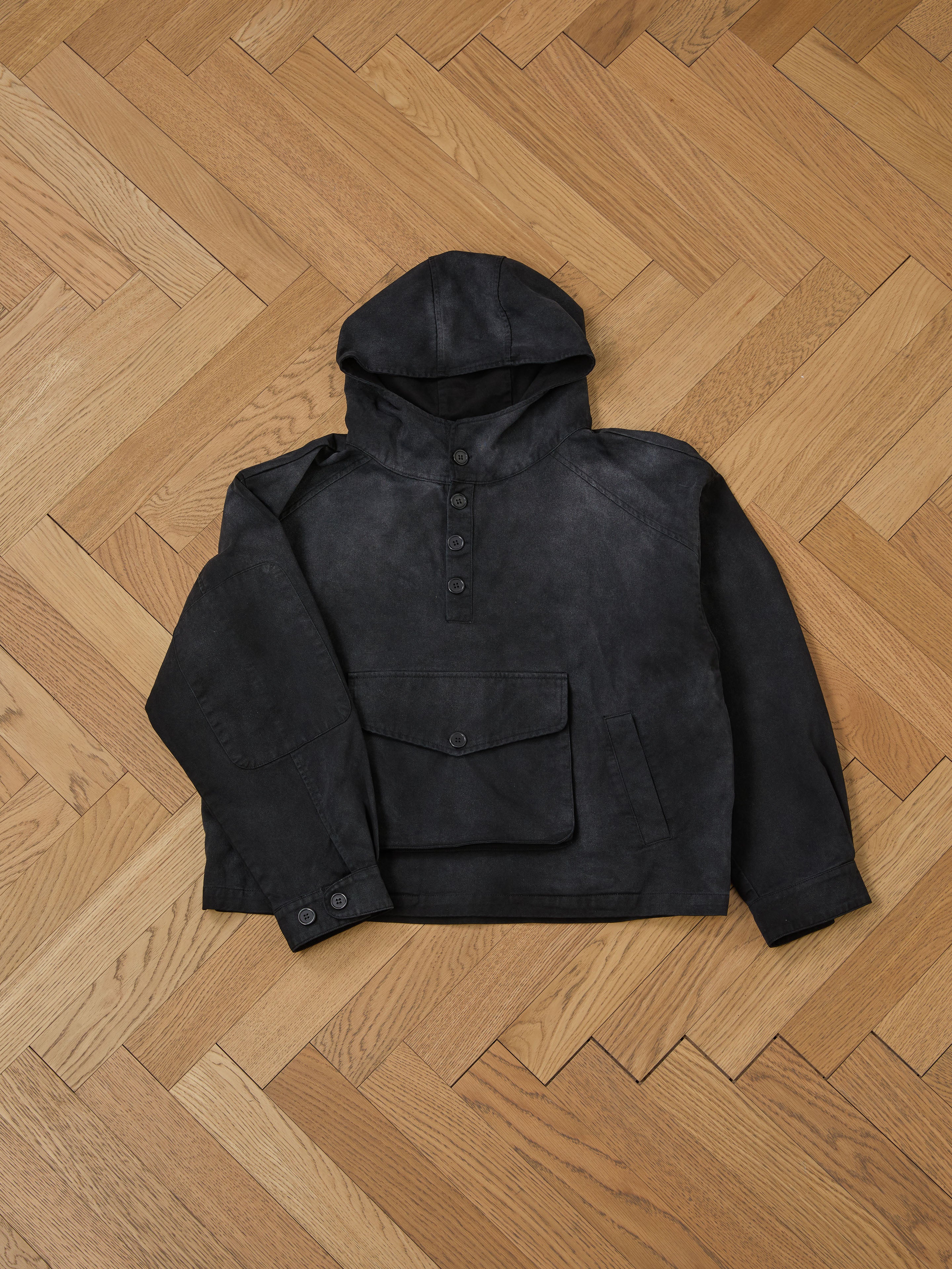 FOUND's Washed Twill Pocket Anorak Jacket, elegantly displayed on a wooden herringbone-patterned floor, is a timeless piece with a dark gray hue, front pocket, buttoned collar, and embodies a unisex relaxed fit.