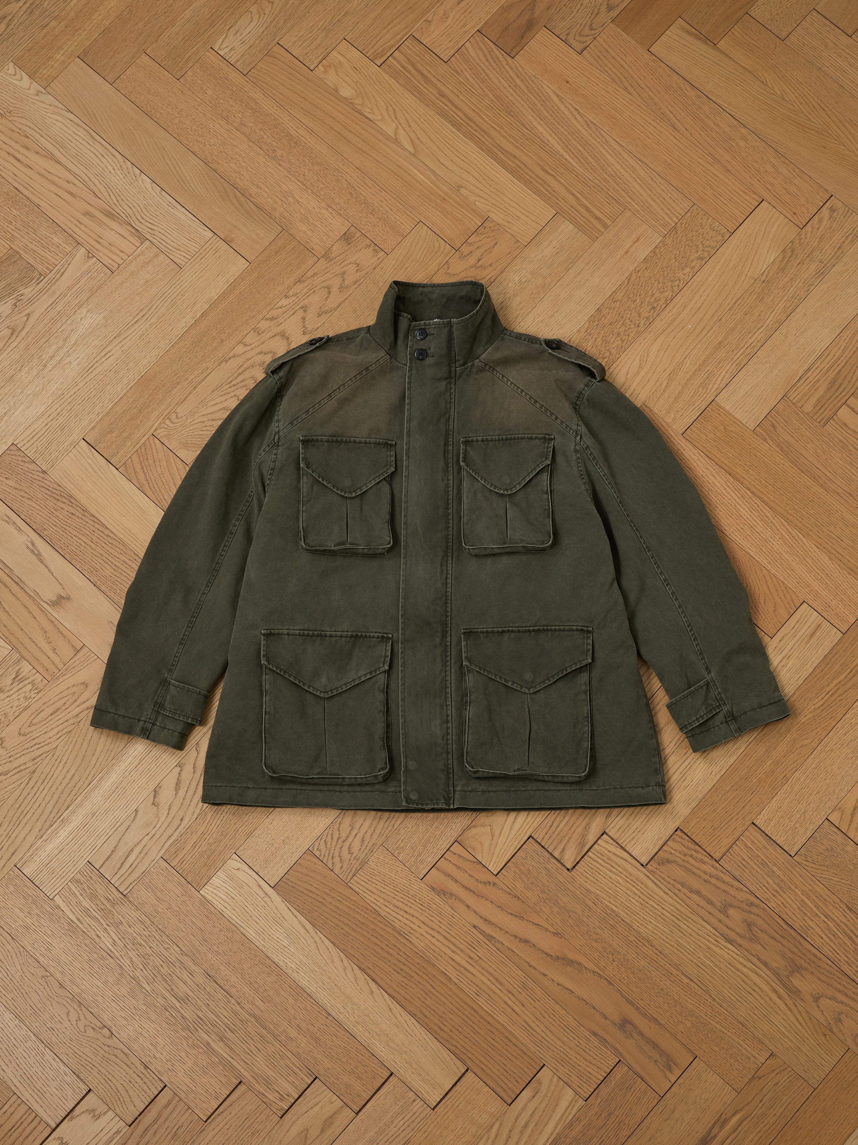 The M65 Utility Twill Pocket Jacket by FOUND is a unisex, military-inspired piece in olive green, featuring four front pockets and a button closure. It's laid flat on a wooden herringbone-patterned floor and combines functionality with timeless appeal in a relaxed fit.