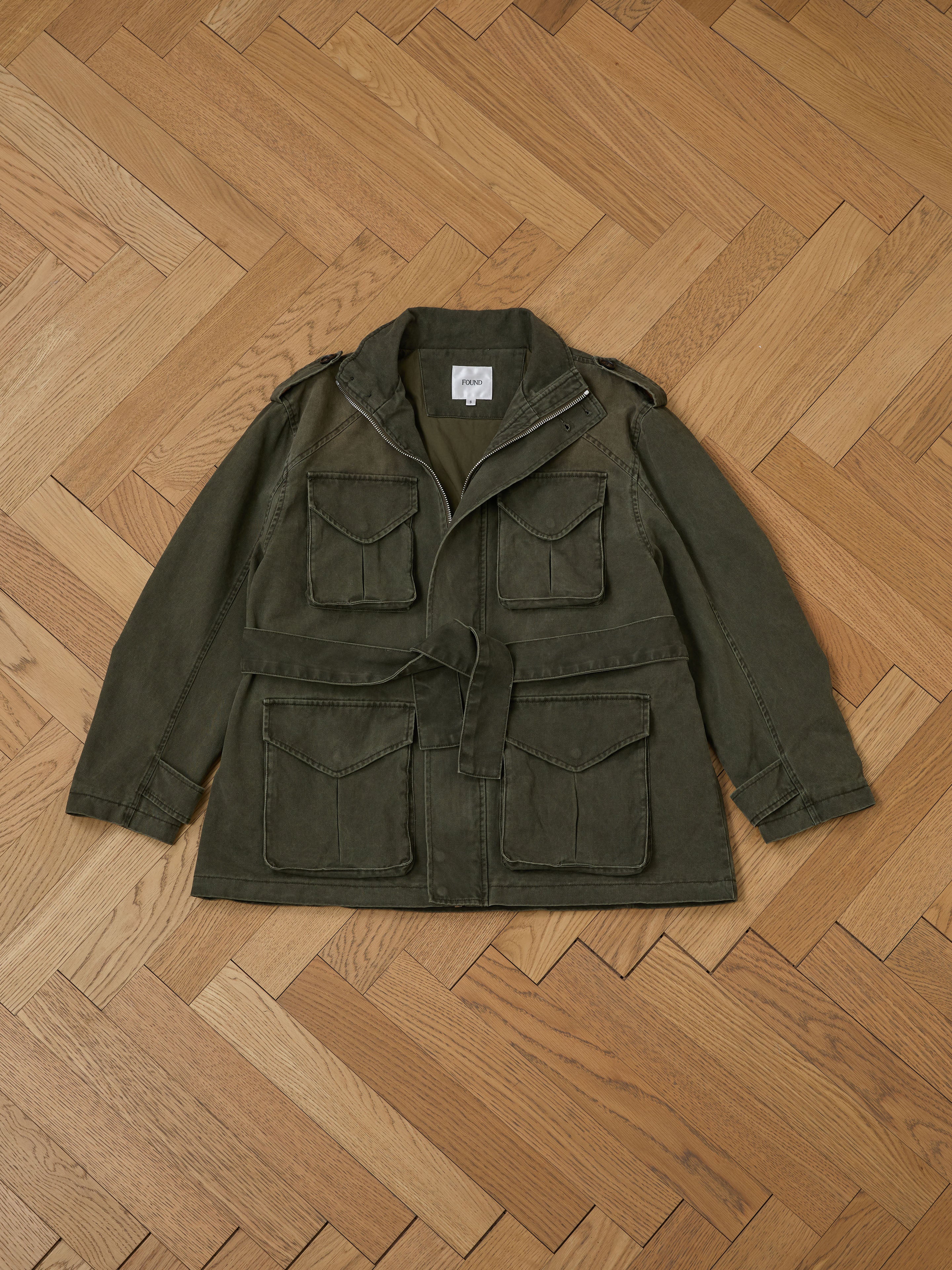 The M65 Utility Twill Pocket Jacket by FOUND features large pockets and a belt, offered in olive green with a military-inspired unisex relaxed fit, stylishly displayed on a wooden herringbone floor.