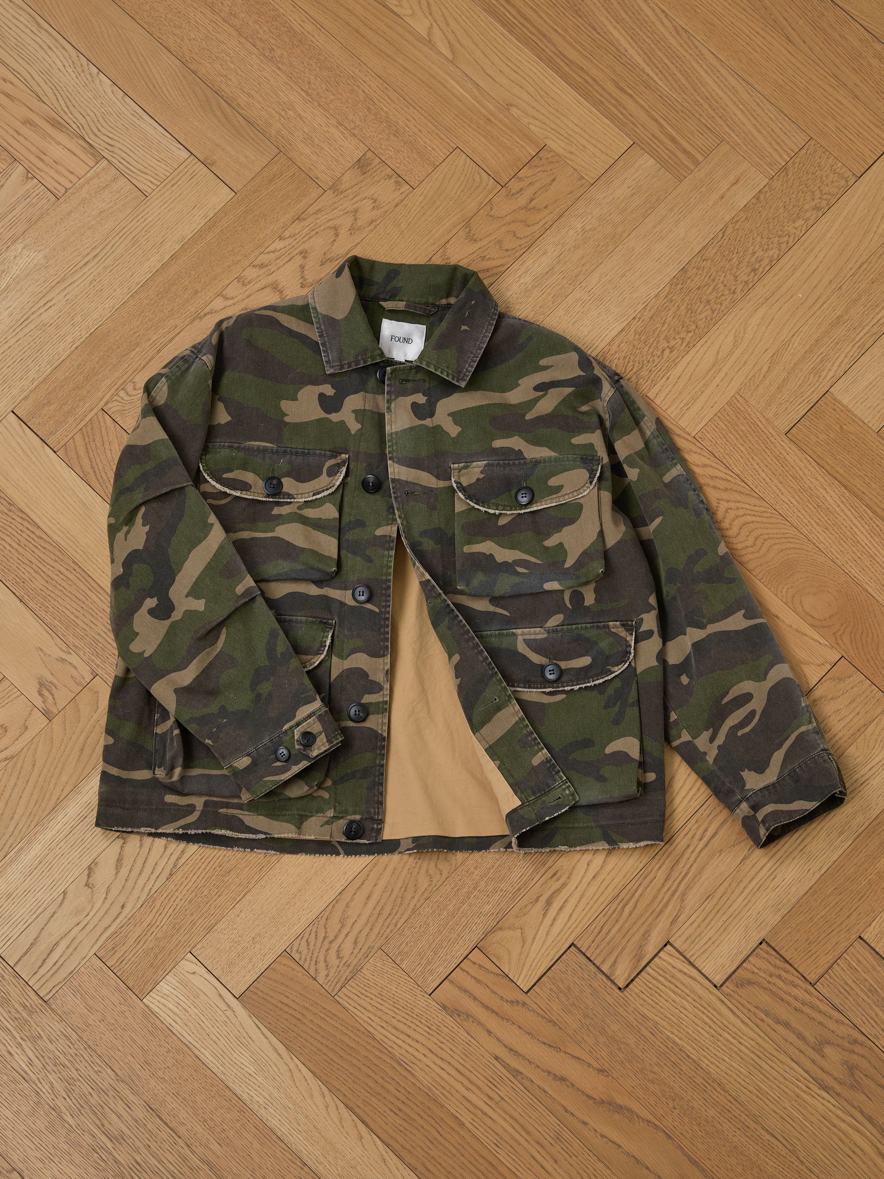 The Camouflage Twill Utility Cargo Jacket by FOUND, with a vintage style, button closure, and two chest pockets, is displayed laid flat on a wooden parquet floor.