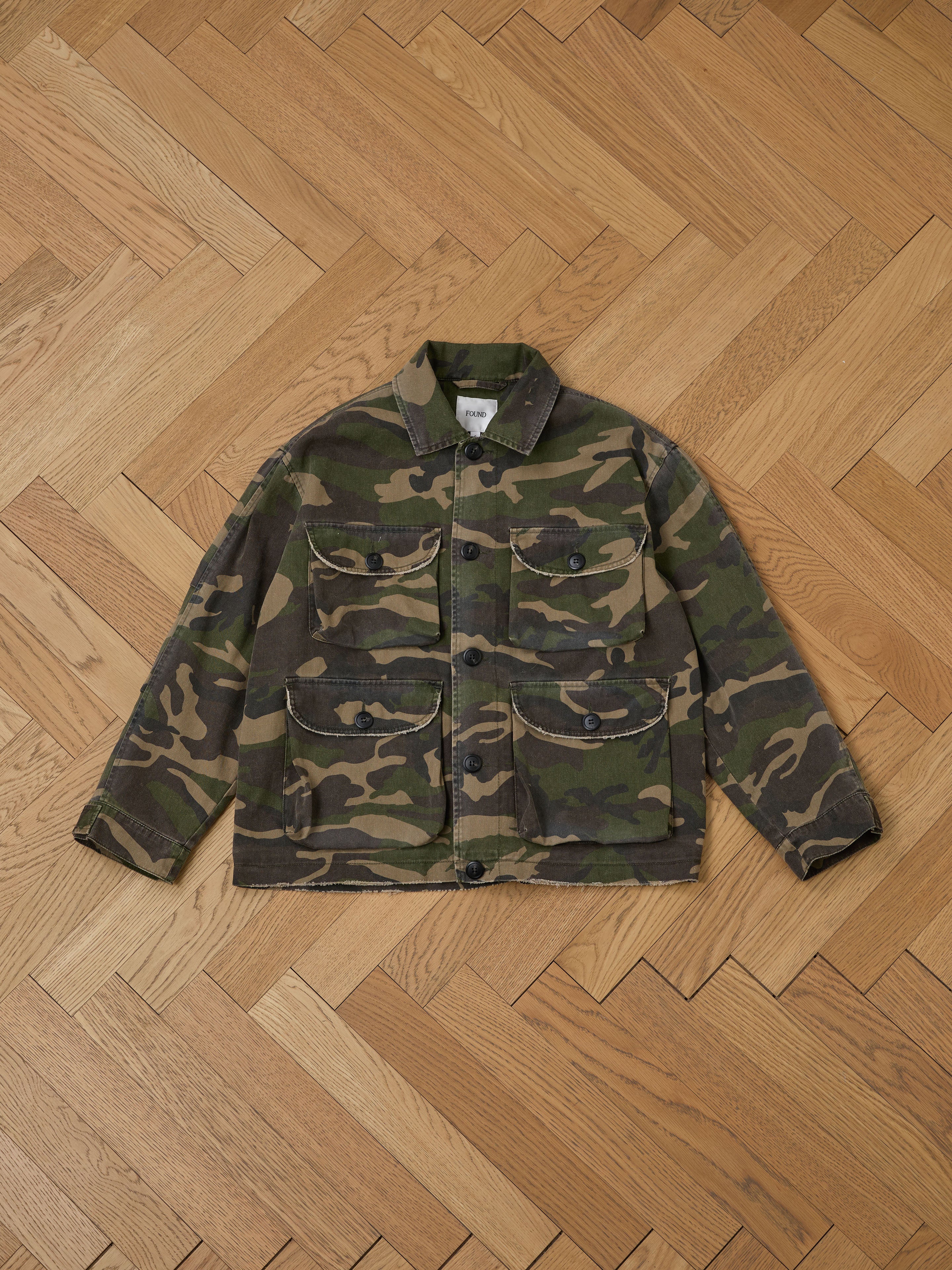 The Camouflage Twill Utility Cargo Jacket by FOUND features a vintage look with four front pockets and button closures, perfect for those who value style and functionality, displayed on a wooden herringbone floor.