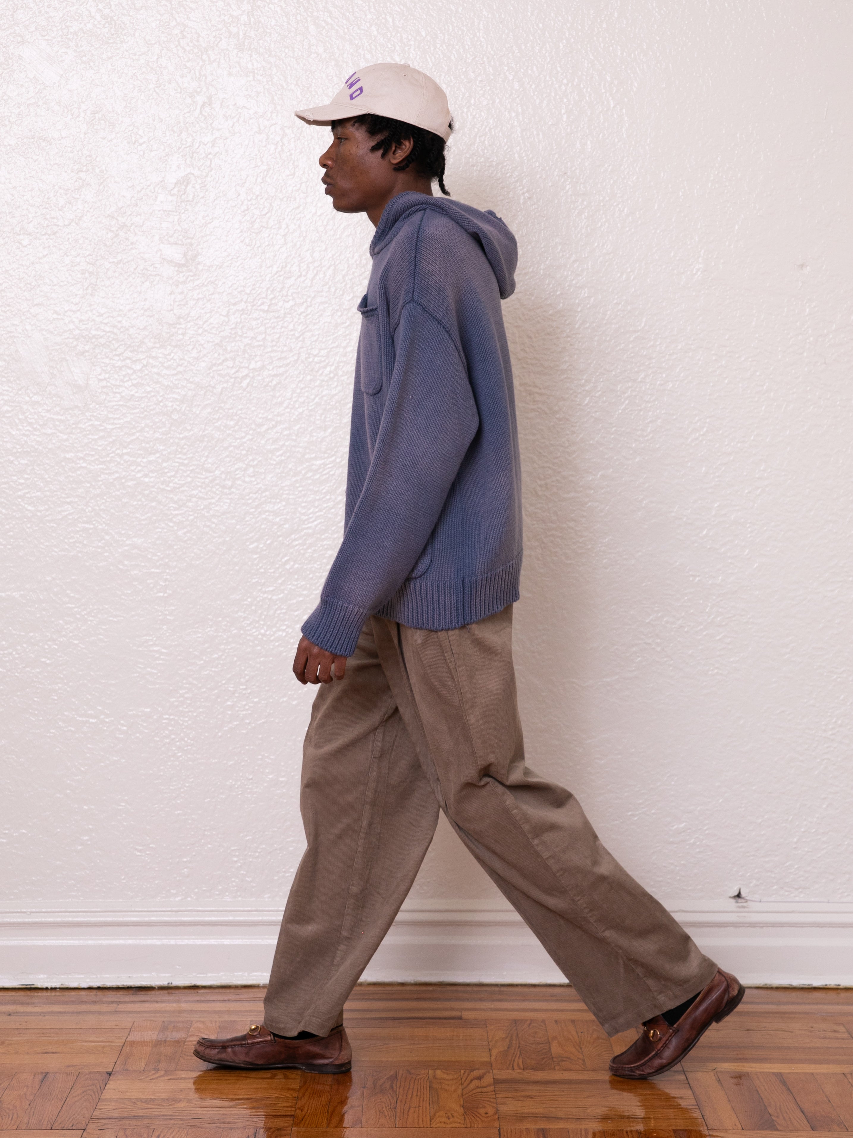 A person in a sand wash-colored Faded Knit Pocket Hoodie by FOUND, beige pants, and a white cap strolls gracefully on a wooden floor against a pristine white wall.