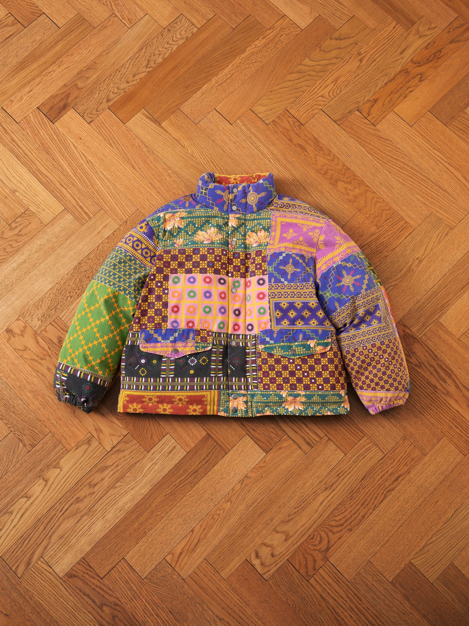 The Ayubia Printed Tapestry Puffer Jacket by FOUND, adorned with various patterns and designs reminiscent of South Asian prints, displayed beautifully on a wooden herringbone floor.