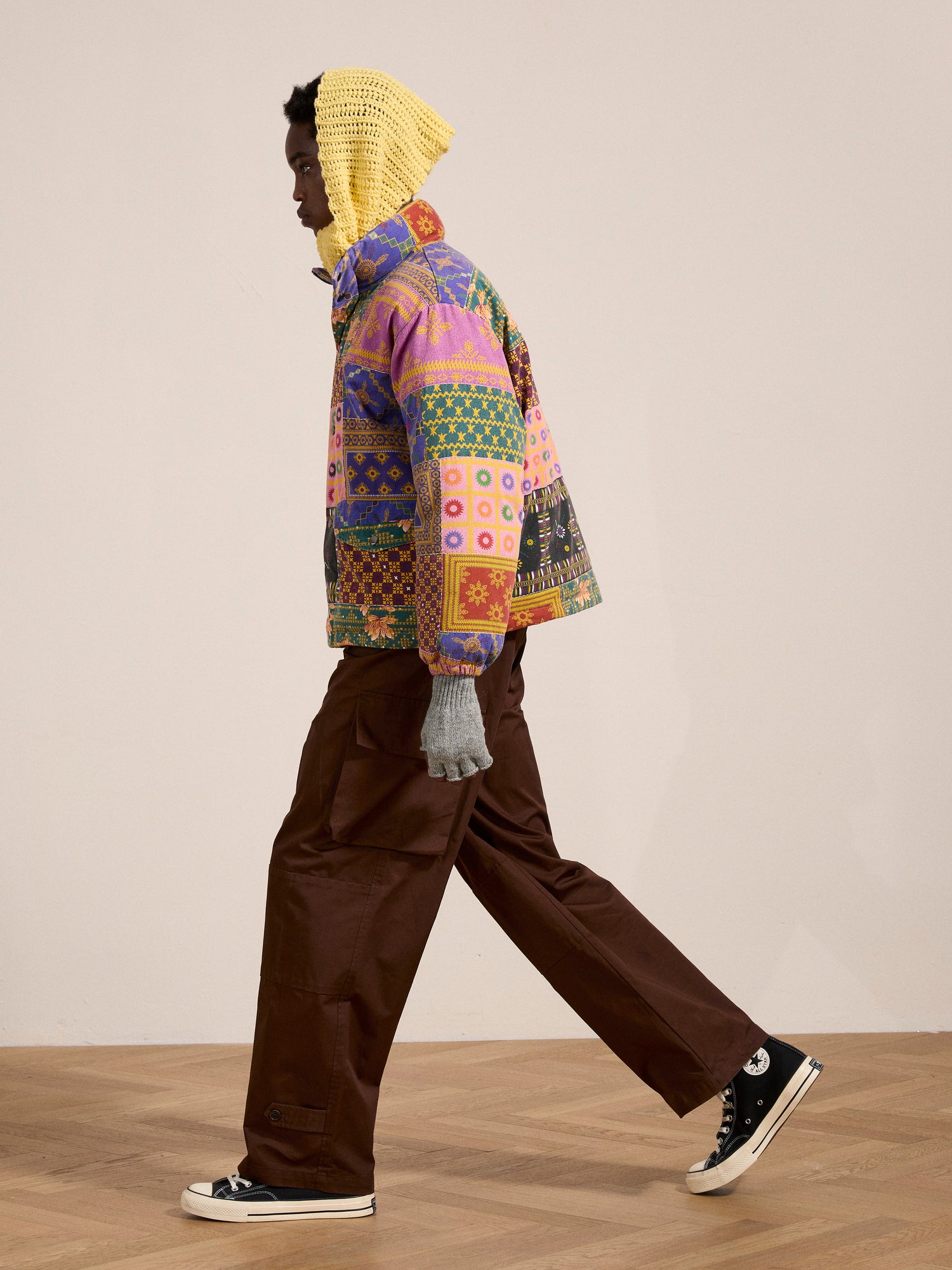 A person wearing the Ayubia Printed Tapestry Puffer Jacket by FOUND along with brown pants, black sneakers, and a yellow knit hood walks across a wooden floor.