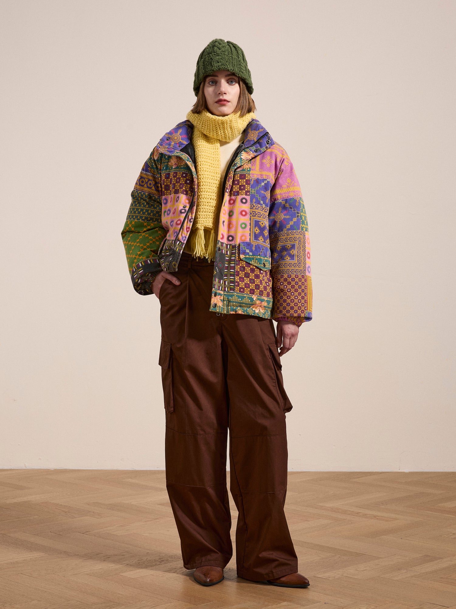 Person wearing a green knit hat, yellow scarf, and the FOUND Ayubia Printed Tapestry Puffer Jacket with vibrant South Asian prints over brown cargo pants, standing on a wooden floor against a plain beige background.