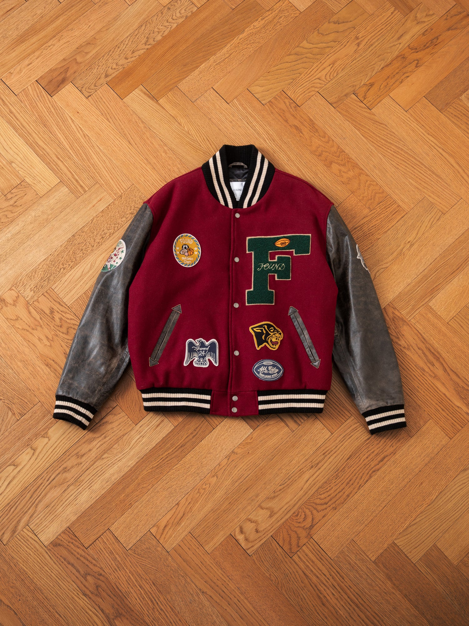 The Crimson Letterman Jacket by Found is laid flat on a wooden herringbone floor. This vintage-look jacket features black leather sleeves and various patches, including a large green 