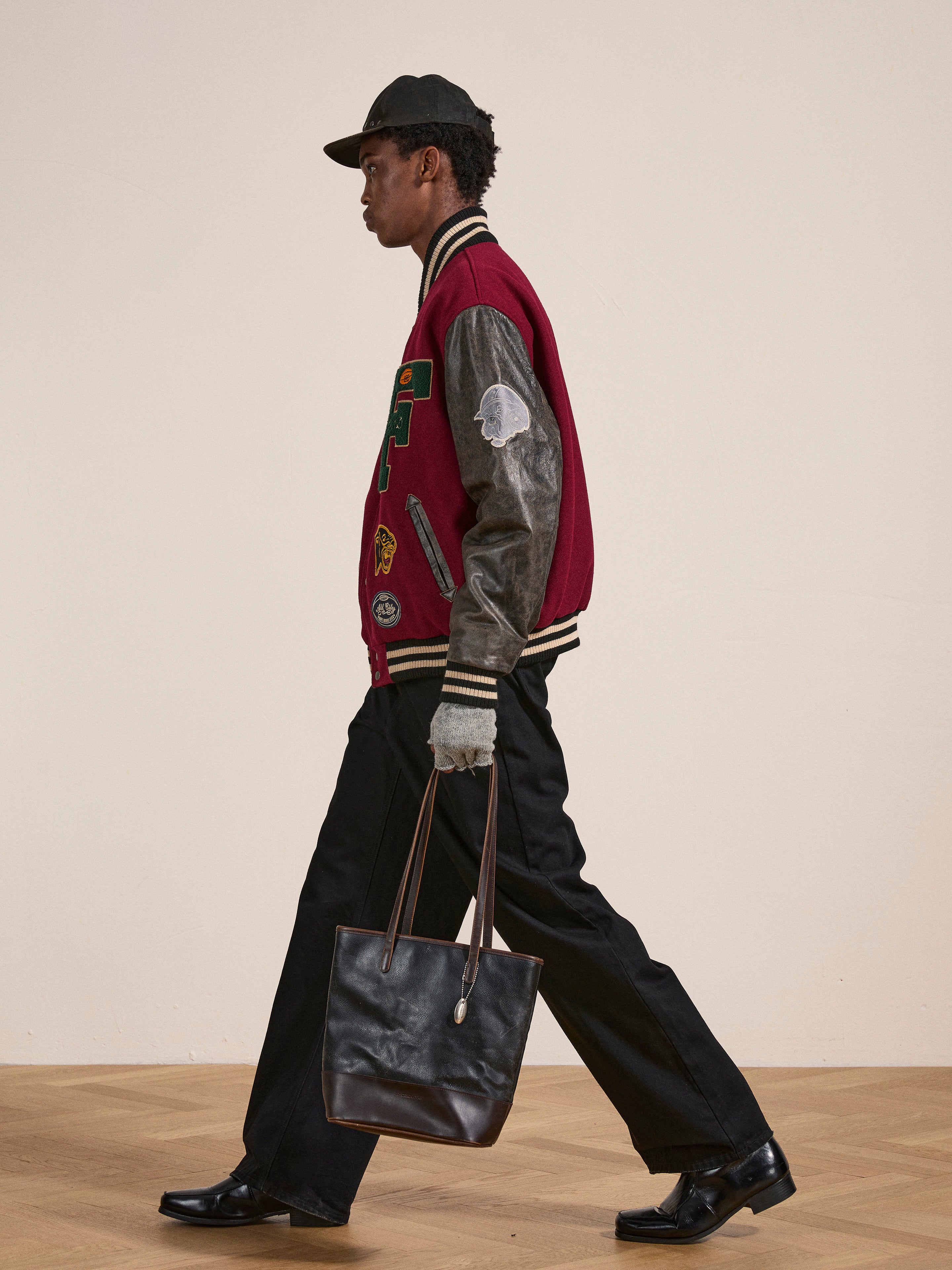 A person wearing a Found Crimson Letterman Jacket, black cap, and black pants walks while holding a black tote bag. The individual is pictured from the side, set against a plain background, showcasing a vintage look.