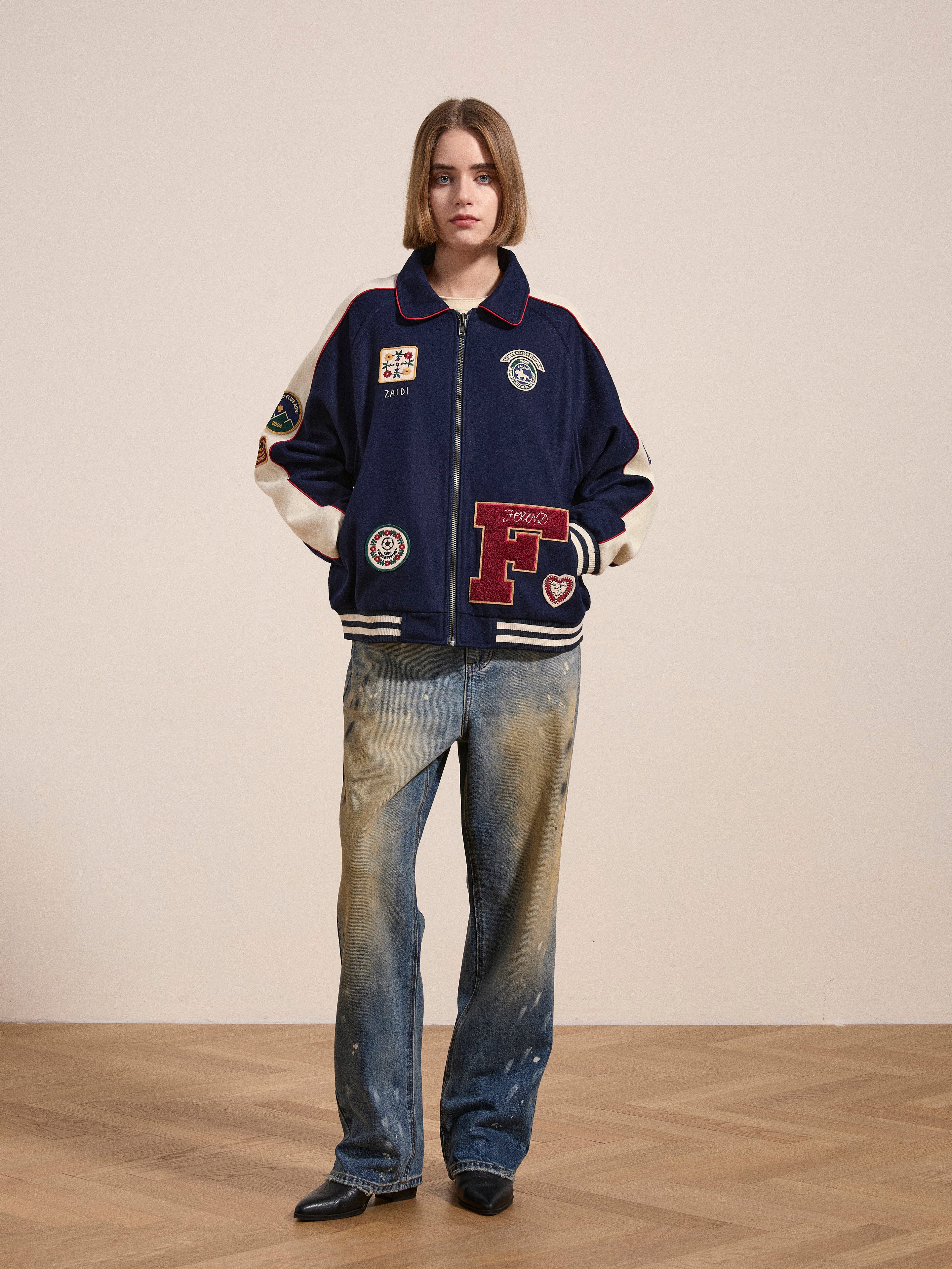 A person stands in front of a plain wall wearing a FOUND All Roads Varsity Jacket with a vintage look, crafted from a mid-weight wool blend, paired with faded, distressed jeans.