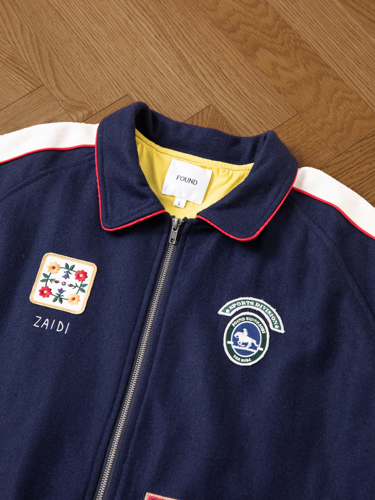 A navy blue All Roads Varsity Jacket from FOUND, featuring a front zipper and adorned with patches such as a floral design, a horse logo, and the name 