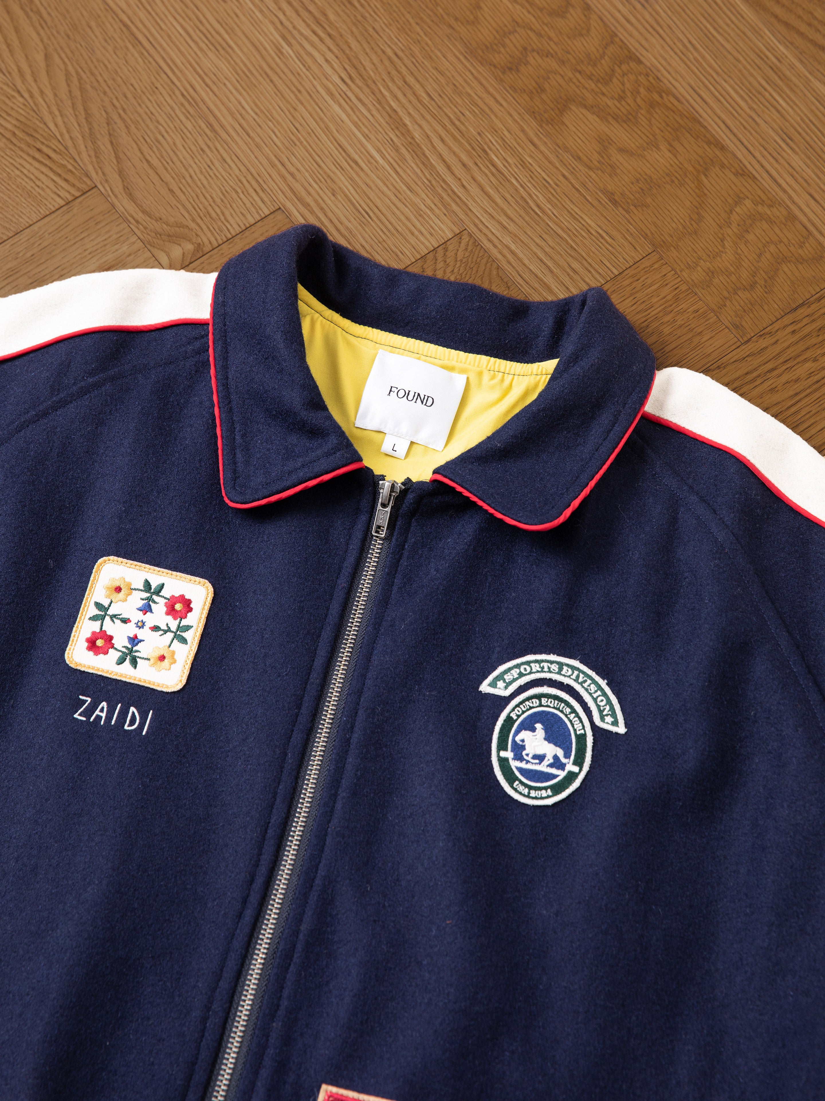 A navy blue All Roads Varsity Jacket from FOUND, featuring a front zipper and adorned with patches such as a floral design, a horse logo, and the name "ZAIDI" on one side, laid on a wooden surface. The mid-weight wool blend fabric provides a vintage aesthetic that is both stylish and durable.