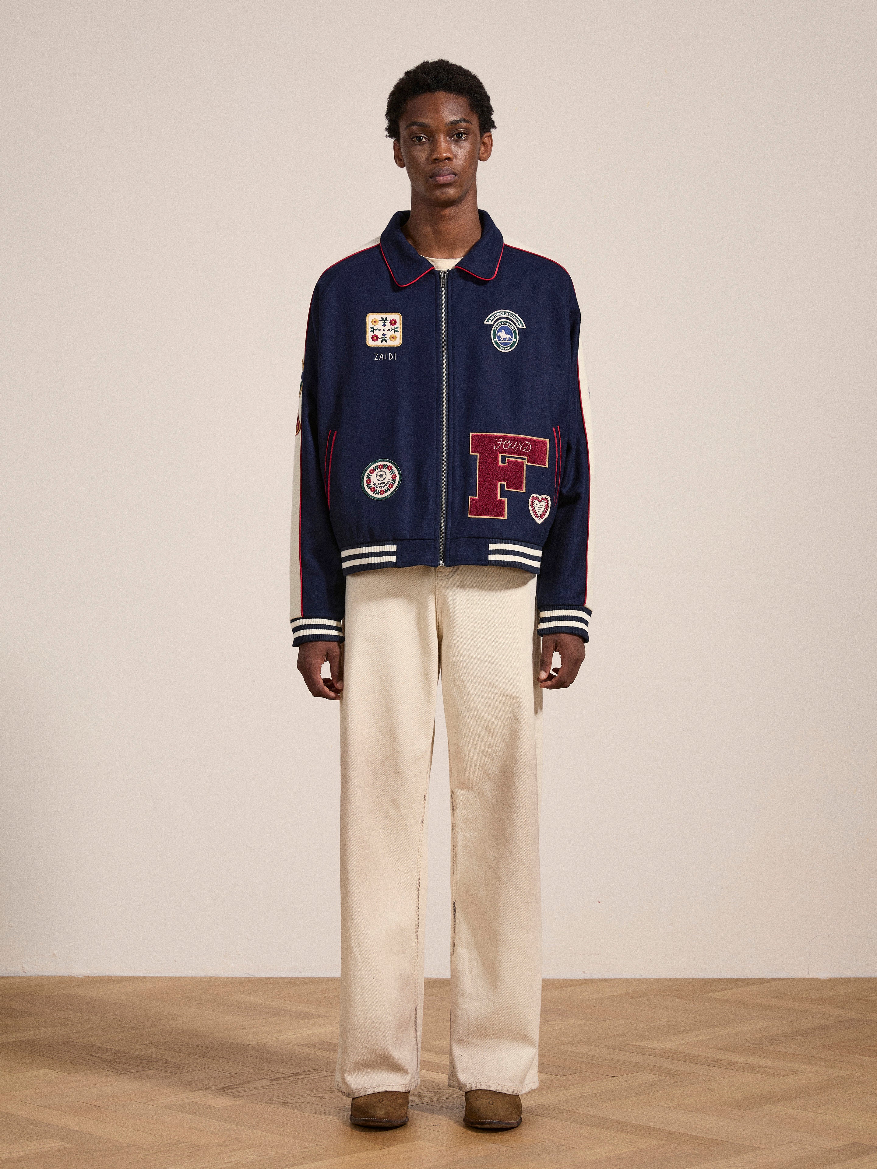 A person stands against a plain background, clad in a navy blue FOUND All Roads Varsity Jacket crafted from a mid-weight wool blend and decorated with patches, paired with cream-colored pants and brown shoes.