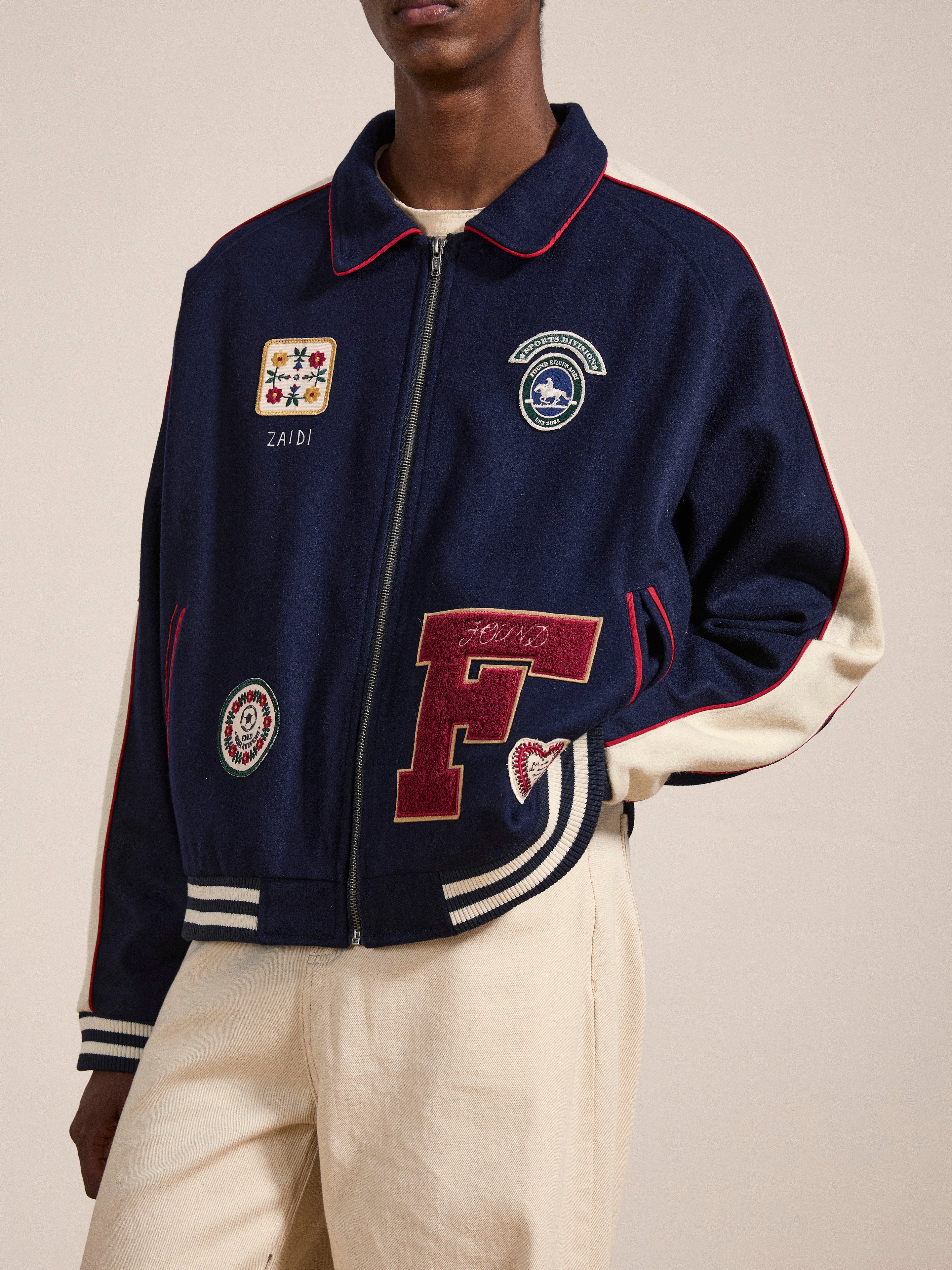 A person is sporting a navy blue All Roads Varsity Jacket by FOUND, complete with embroidered patches, paired with cream-colored pants. This mid-weight wool blend jacket features a prominent red "F" patch along with other smaller patches on the front, contributing to its vintage aesthetic.