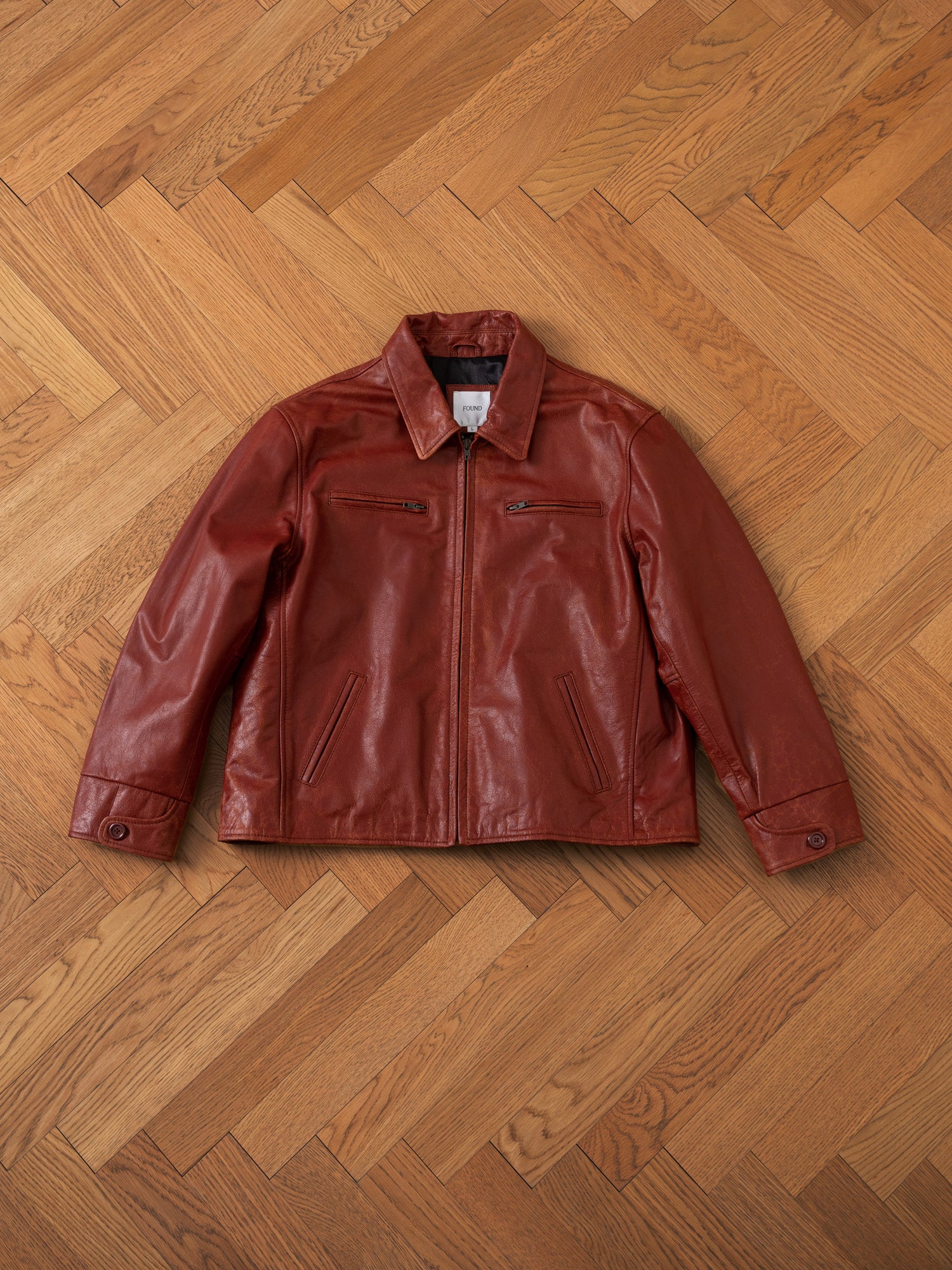 A FOUND Marlboro Sun Faded Leather Racer Jacket, made from ethically sourced materials, with a front zipper and two vertical zippered chest pockets, is laid out flat on a herringbone wooden floor.