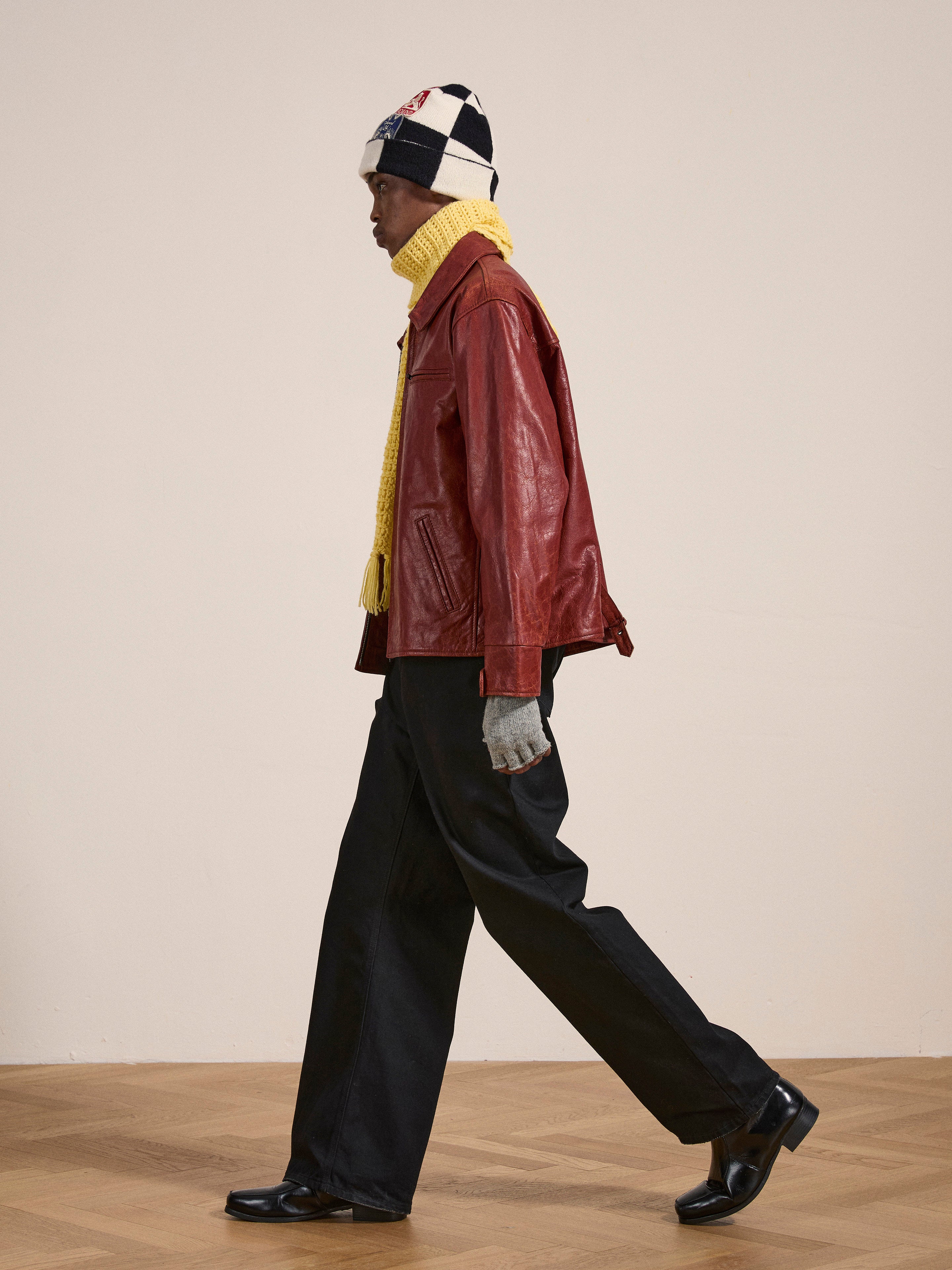 Person wearing a Marlboro Sun Faded Leather Racer Jacket from FOUND, yellow scarf, gray gloves, black pants, black shoes, and a white and black beanie, walking indoors on a wooden floor.