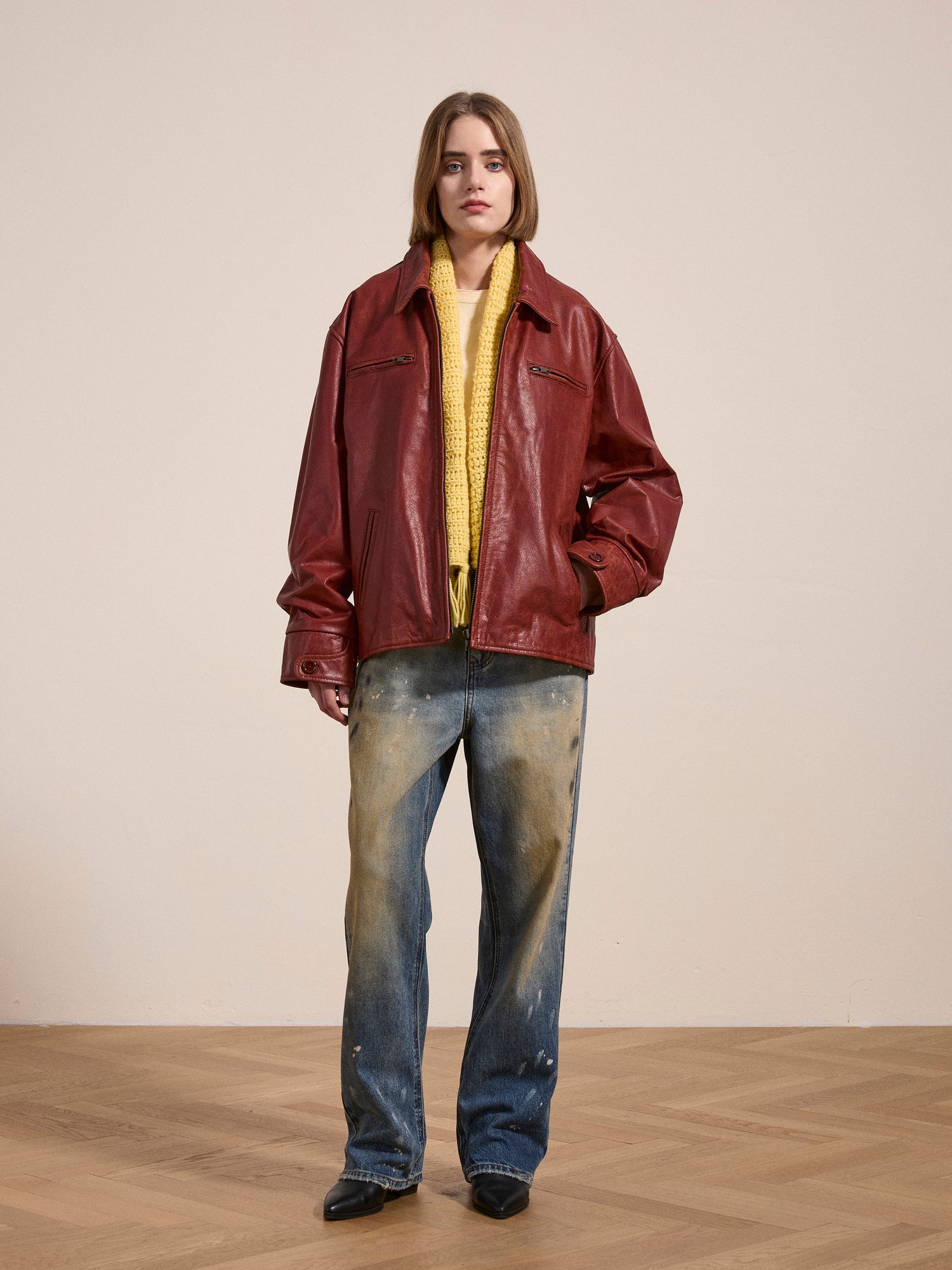 A person stands in a room wearing a Marlboro Sun Faded Leather Racer Jacket by FOUND, a yellow sweater, distressed jeans, and black shoes.