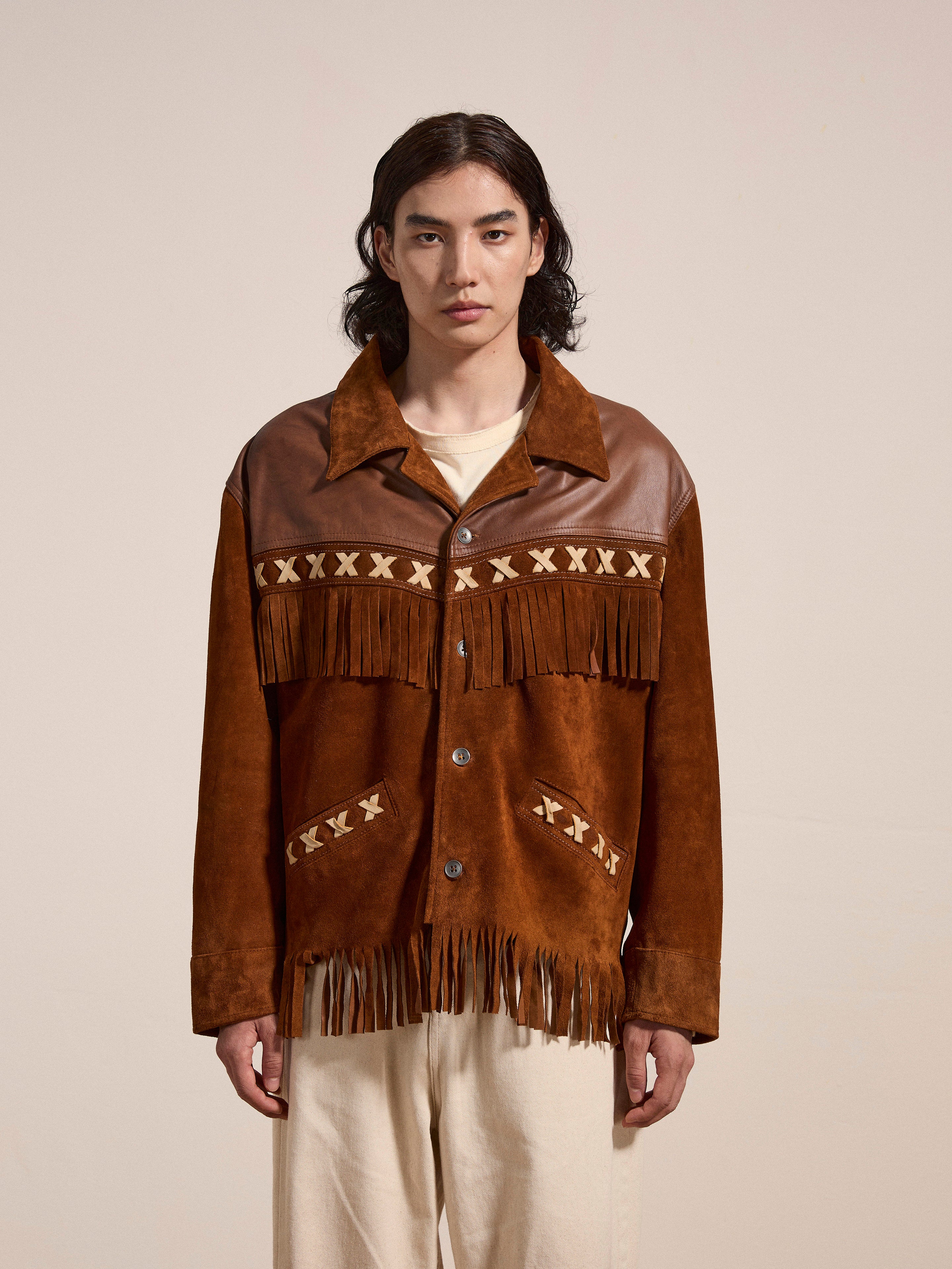 A person with long dark hair stands facing the camera, wearing the Western Fringe Suede Jacket by FOUND, crafted from ethically sourced materials. The brown two-tone suede and leather jacket is adorned with decorative patterns across the chest and pocket areas. They are also wearing light-colored pants.