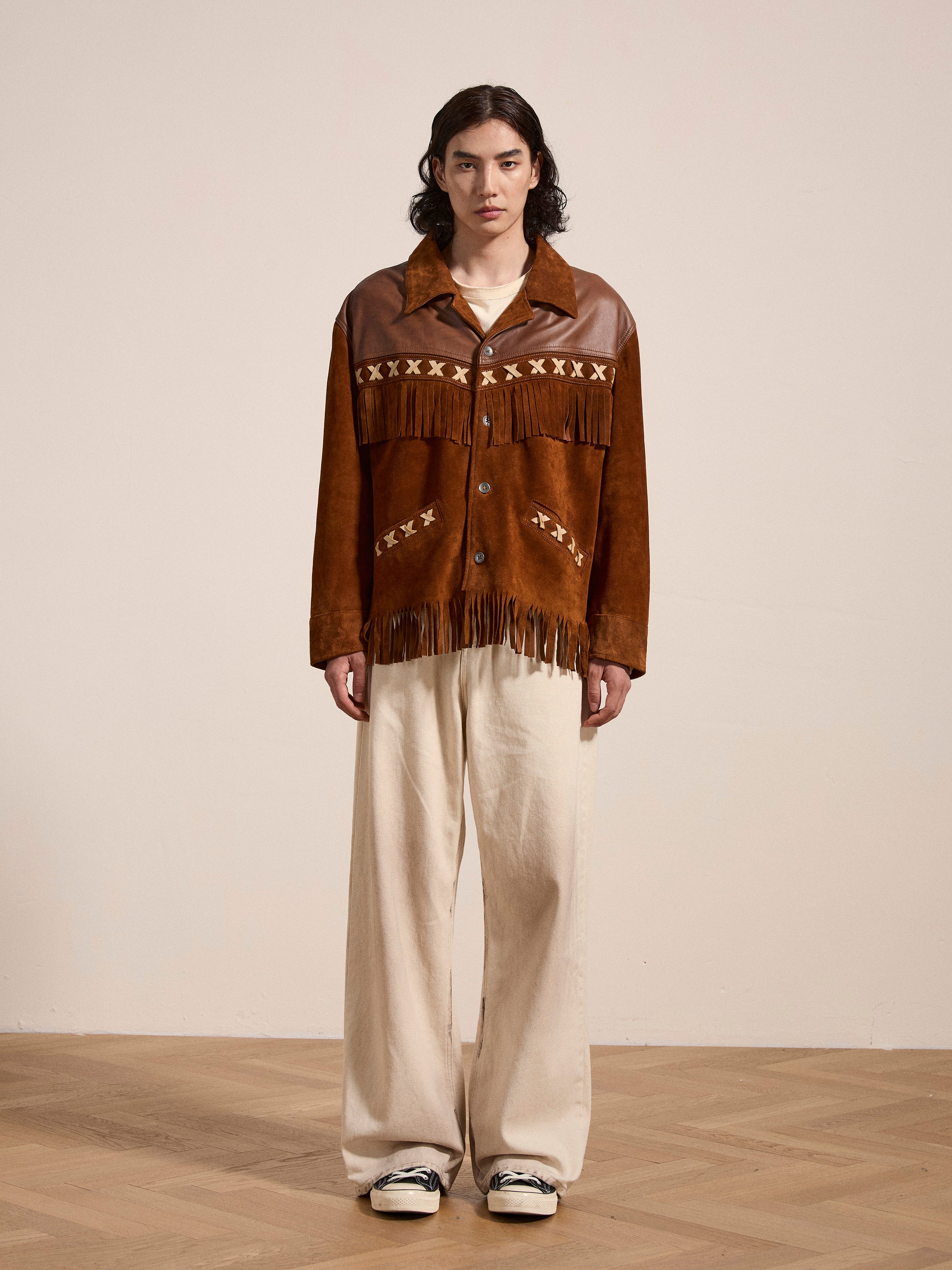 A person stands indoors wearing the Western Fringe Suede Jacket by FOUND, made from ethically sourced materials, paired with beige trousers on a wooden floor.