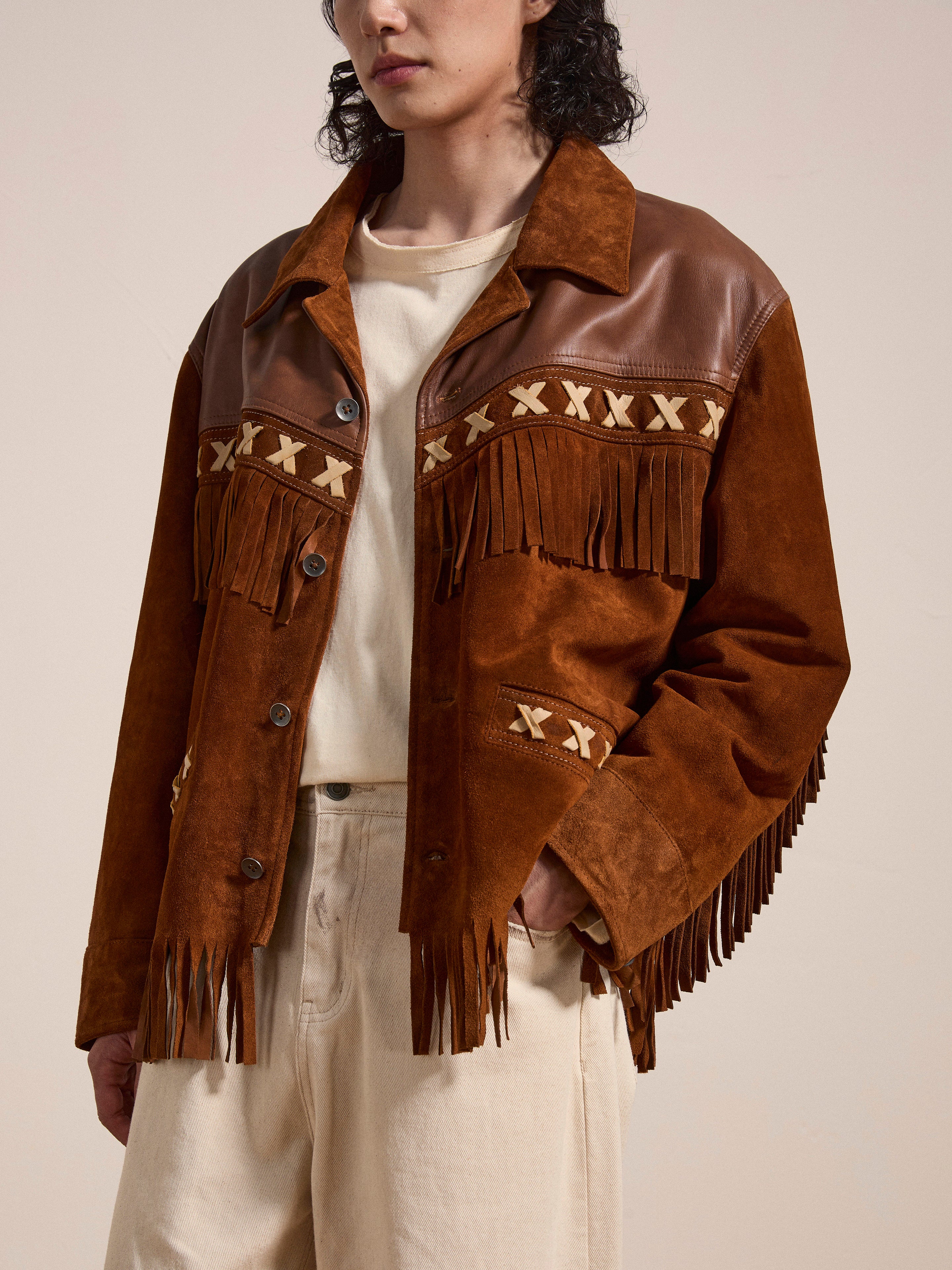 Someone is seen wearing the Western Fringe Suede Jacket by FOUND, adorned with 'X' patterns and crafted from ethically sourced materials. The brown suede jacket features fringe detailing and is paired with a beige shirt and pants, standing against a plain backdrop.