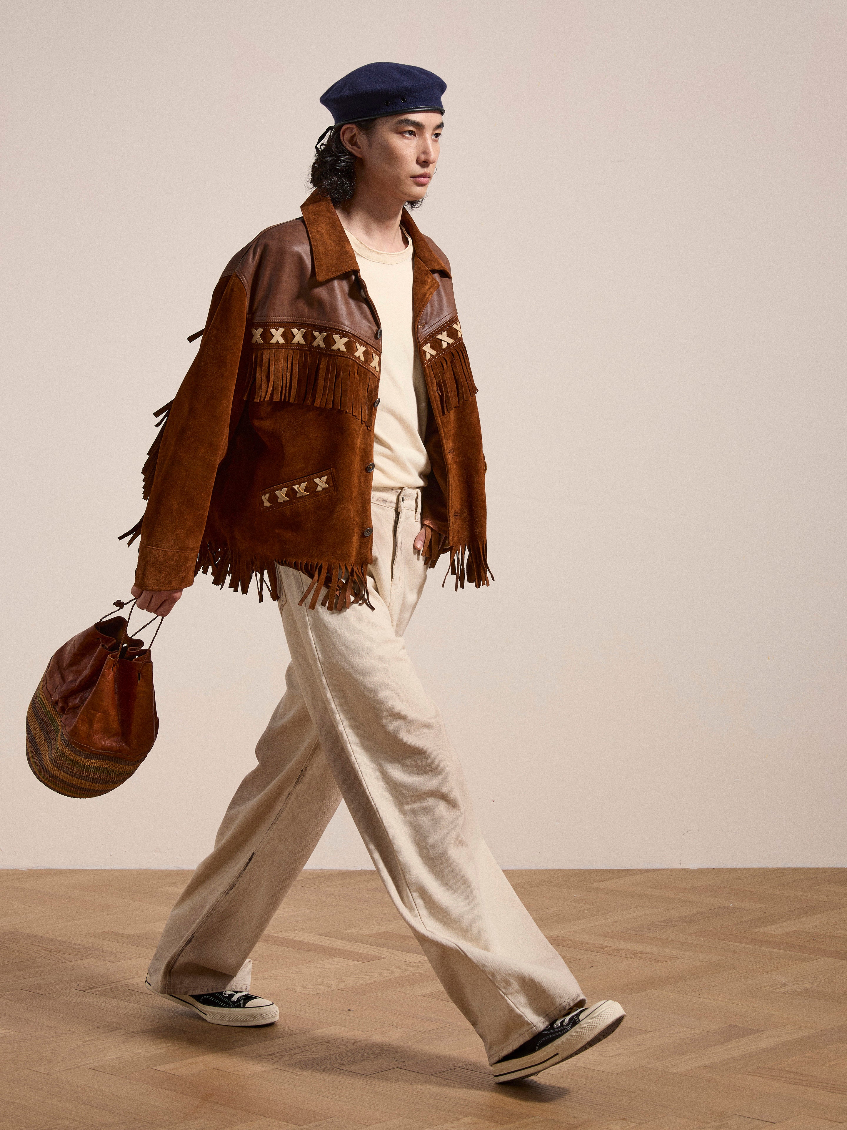 A person walks while carrying a brown bag. They are dressed in the Western Fringe Suede Jacket by FOUND, which is made from ethically sourced materials, along with white pants, black shoes, and a blue beret. The background is plain and minimal.