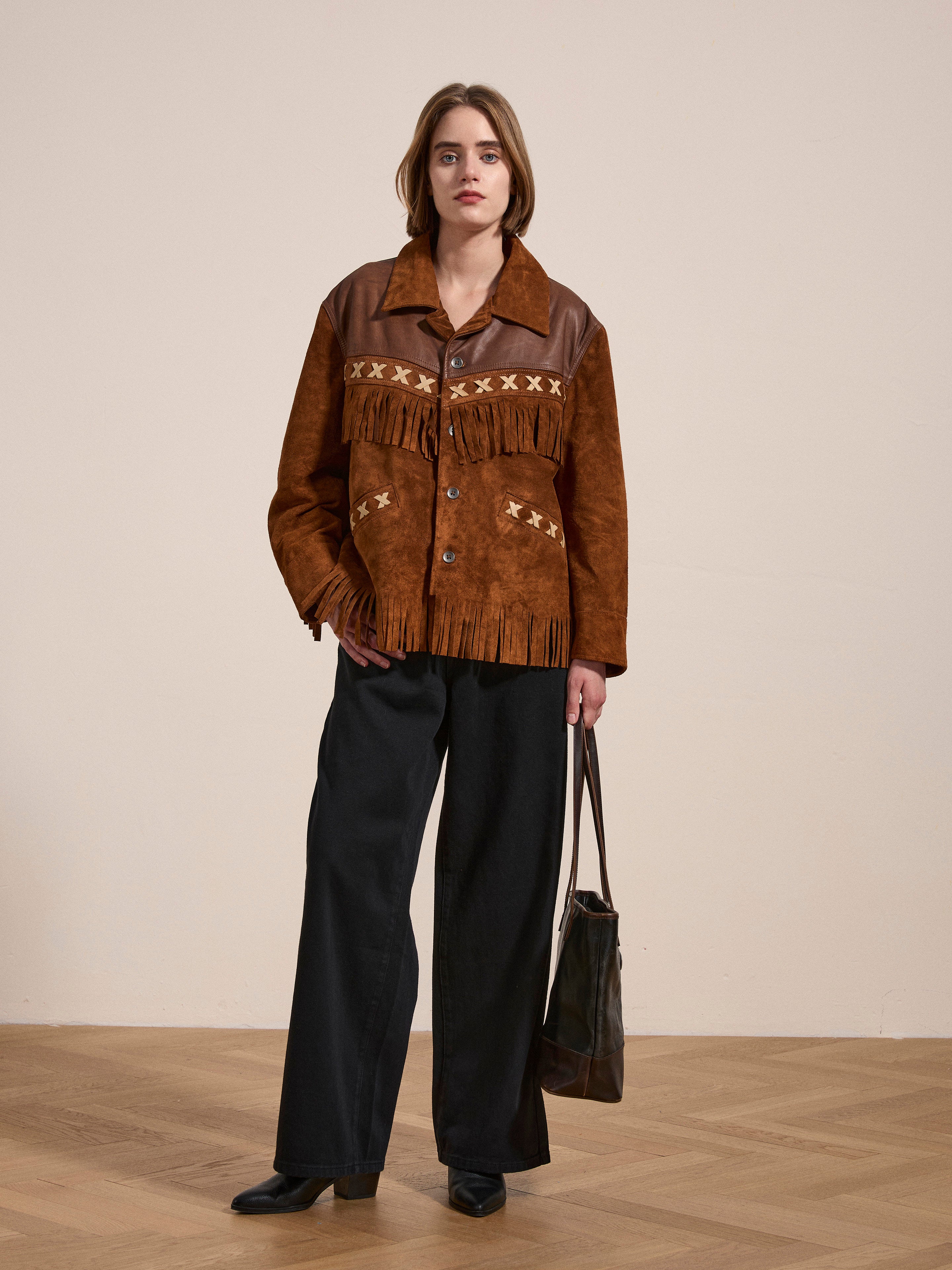 A person stands indoors wearing a Western Fringe Suede Jacket by FOUND, made from ethically sourced materials. They pair it with black wide-leg pants and hold a black handbag against a plain wall backdrop with a wooden floor.