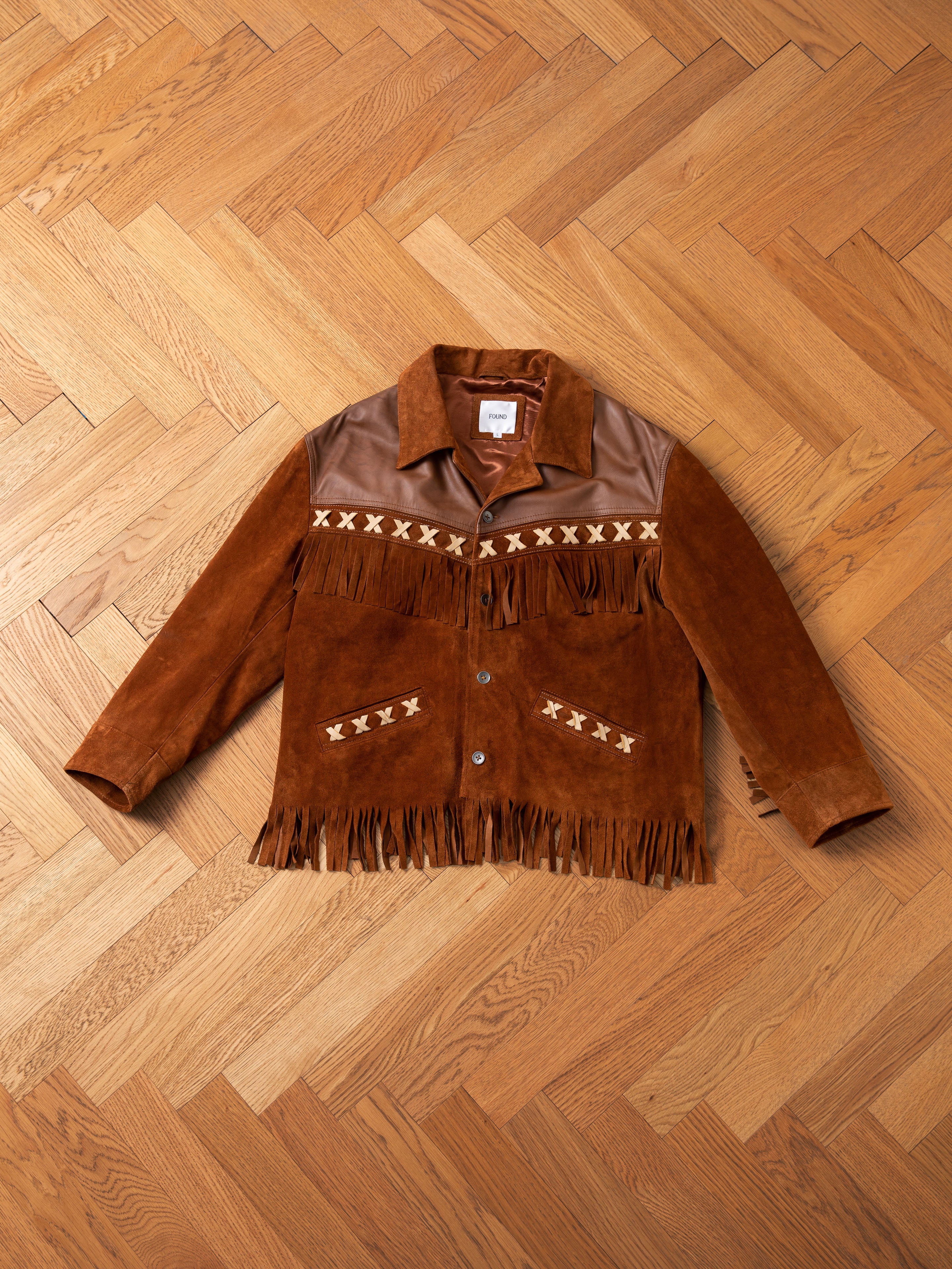 A FOUND Western Fringe Suede Jacket, crafted from ethically sourced materials and showcasing fringe detailing along with geometric patterns across the chest. The button-up front and two-tone suede and leather design are artfully displayed flat on a wooden floor.