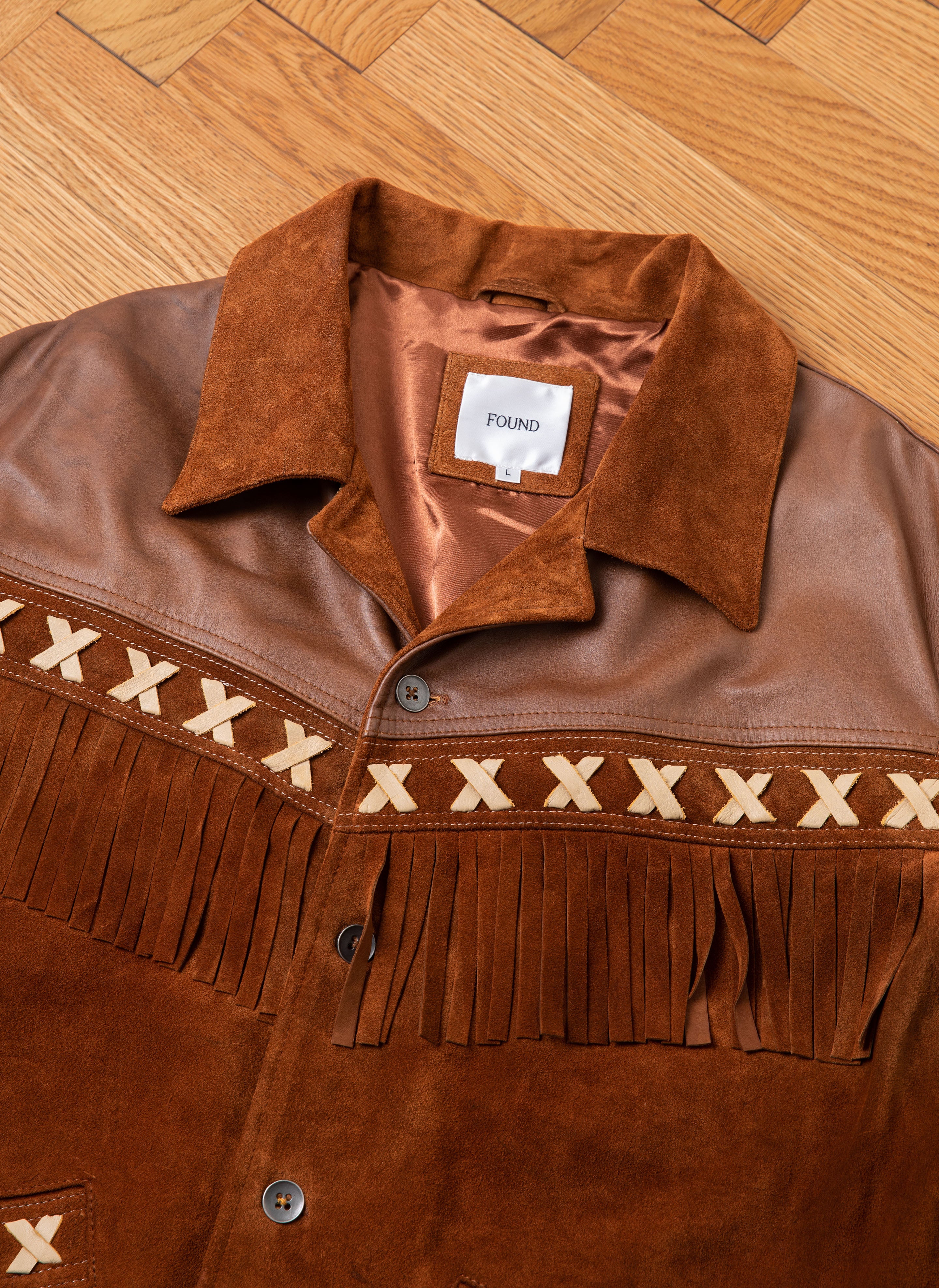 A Western Fringe Suede Jacket, crafted from two-tone suede and leather with decorative fringe and X-pattern details, lies on a wooden floor. The label inside the collar reads "FOUND," proudly indicating its ethically sourced materials.