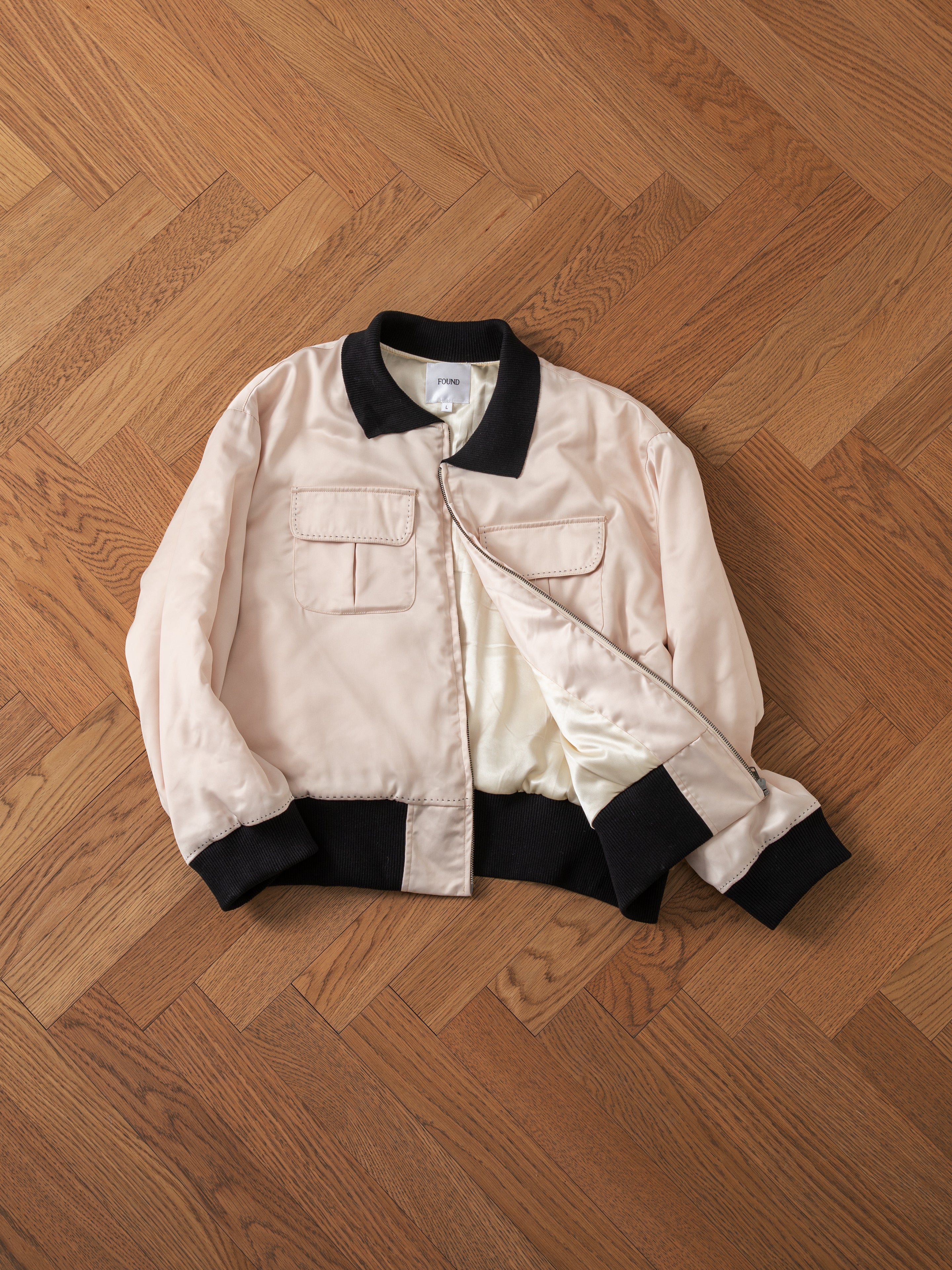 A Daman Satin Jacket by FOUND, featuring a light color with contrasting black collar, cuffs, and waistband, is laid flat on a wooden parquet floor. The jacket also includes front pockets with flaps.