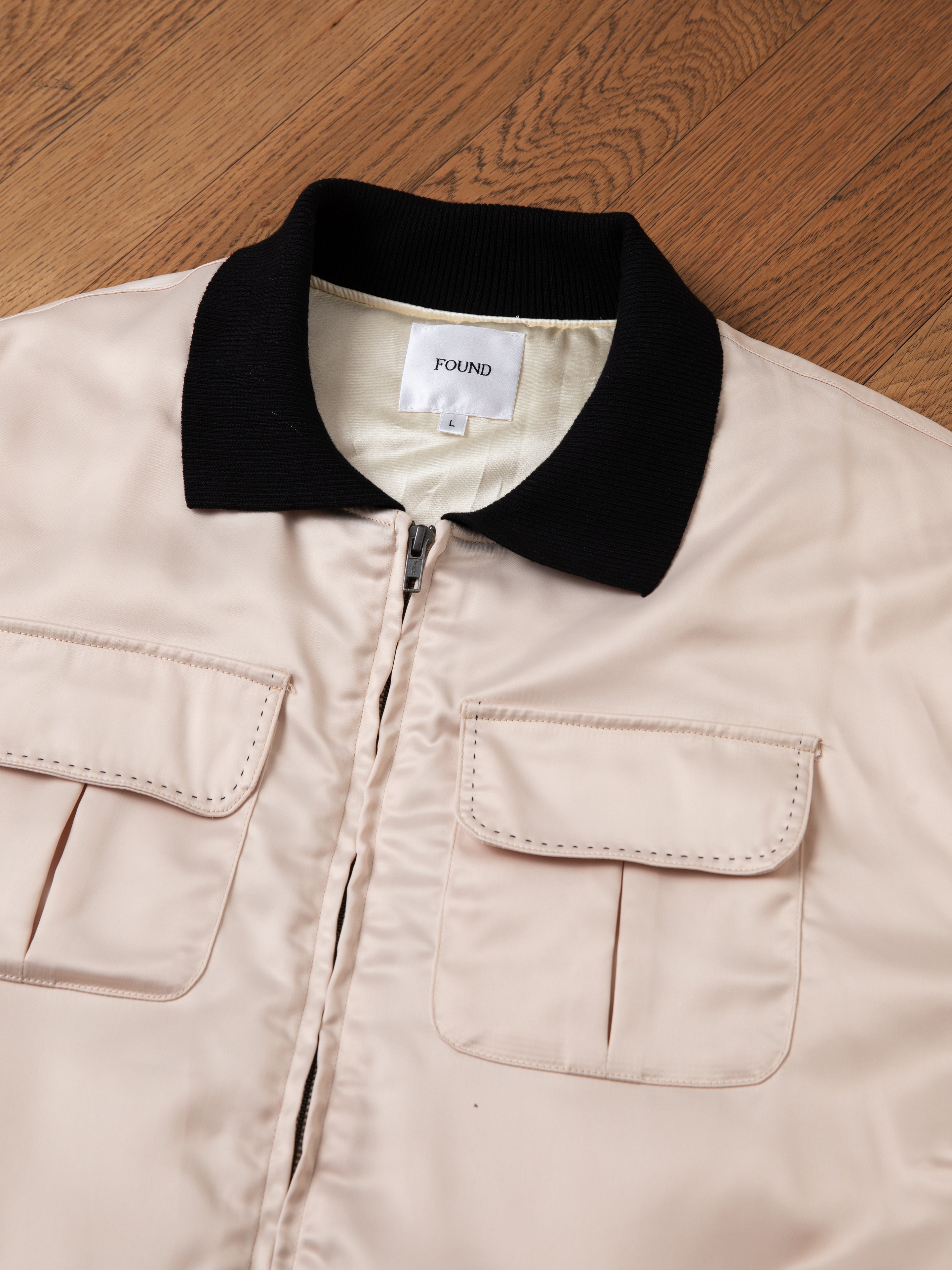 A beige Daman Satin Jacket with a black collar and two front pockets lies flat on a wooden floor. The jacket has a tag labeled "FOUND.