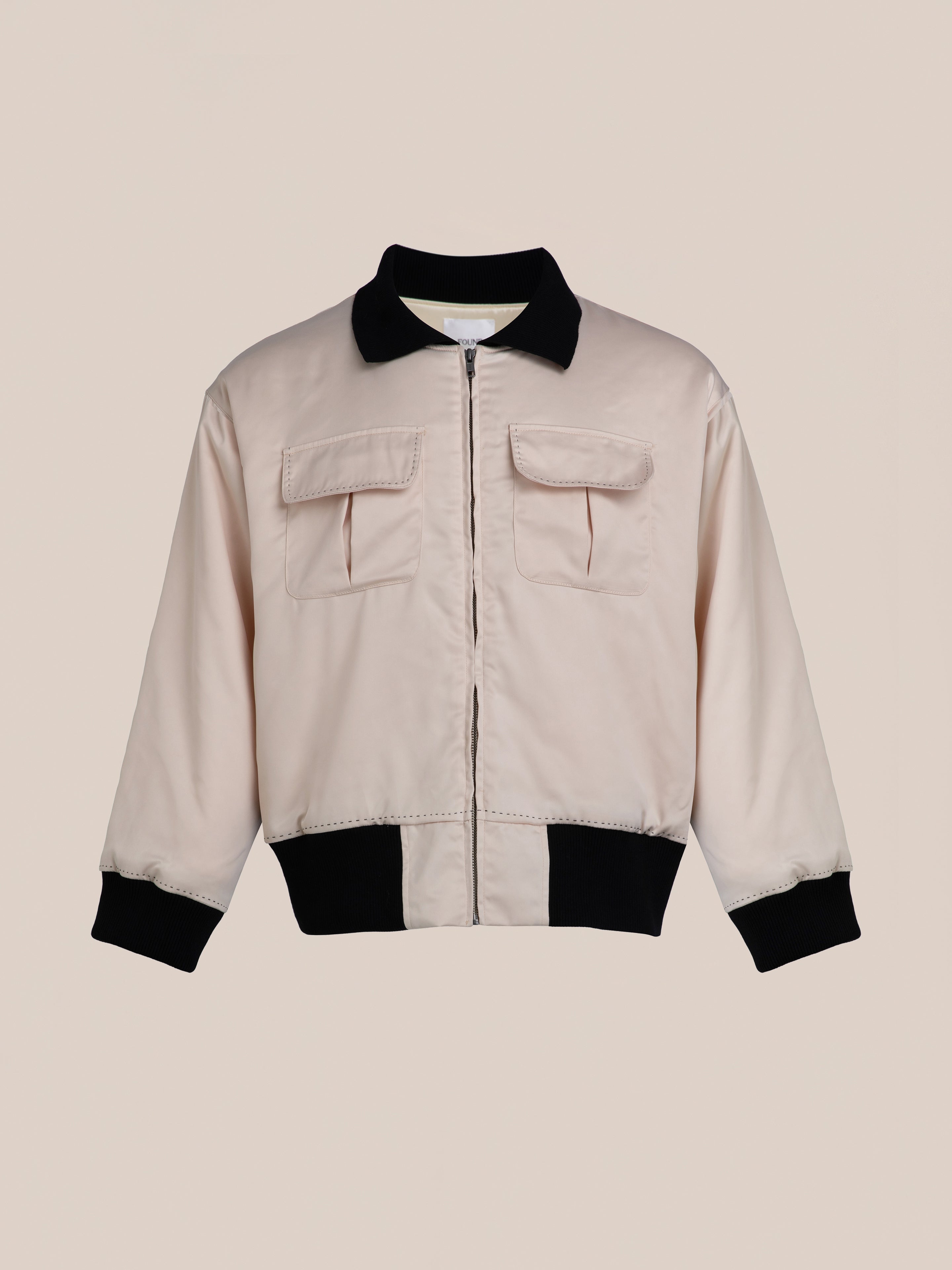 The Daman Satin Jacket by FOUND is a light pink bomber jacket featuring a black collar, black cuffs, and a black hem. It includes two front flap pockets and is designed with a front zipper.