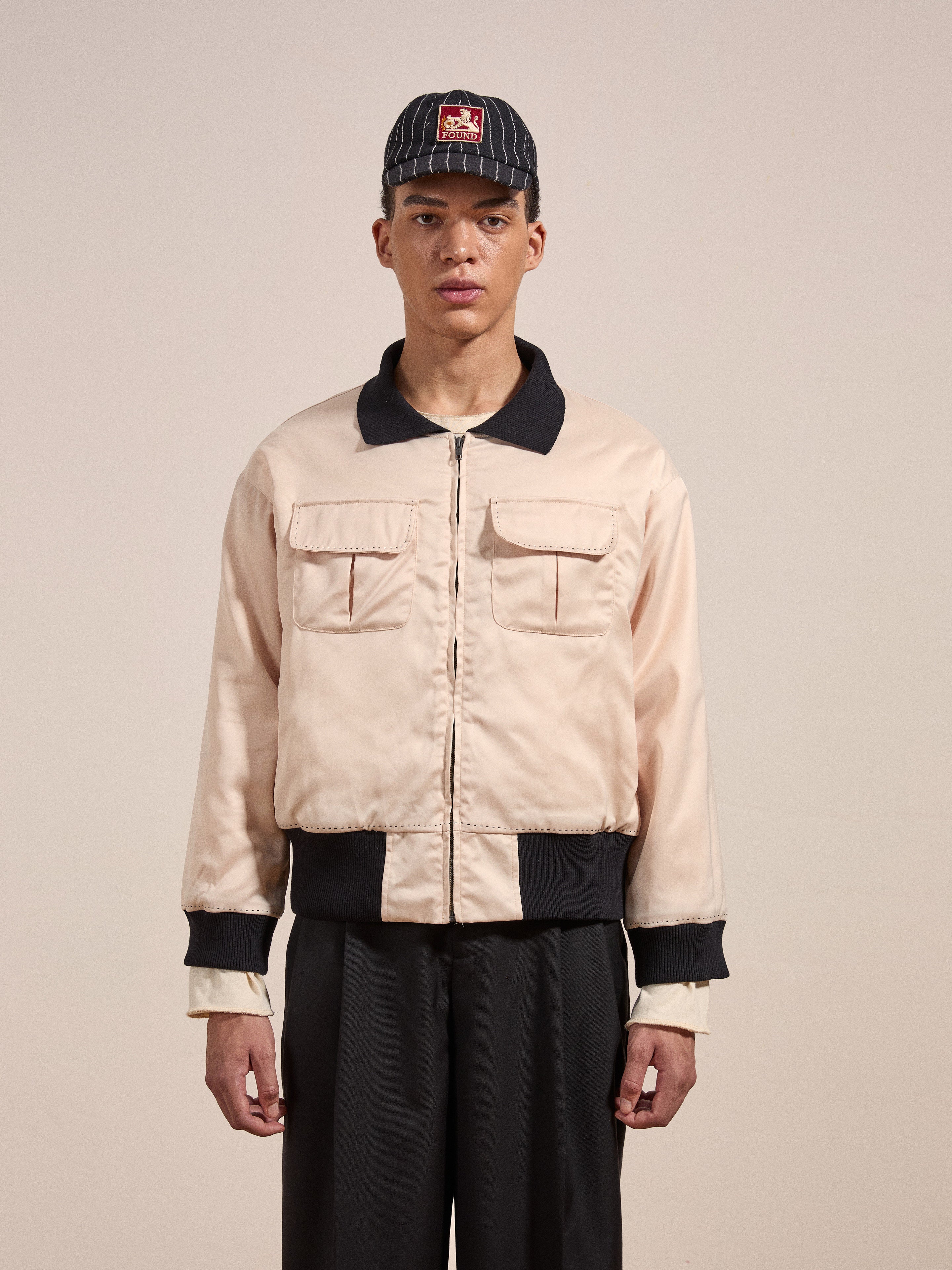 A person wearing the "Daman Satin Jacket" by FOUND, which features a beige color with black trim, and a black baseball cap stands against a plain background.