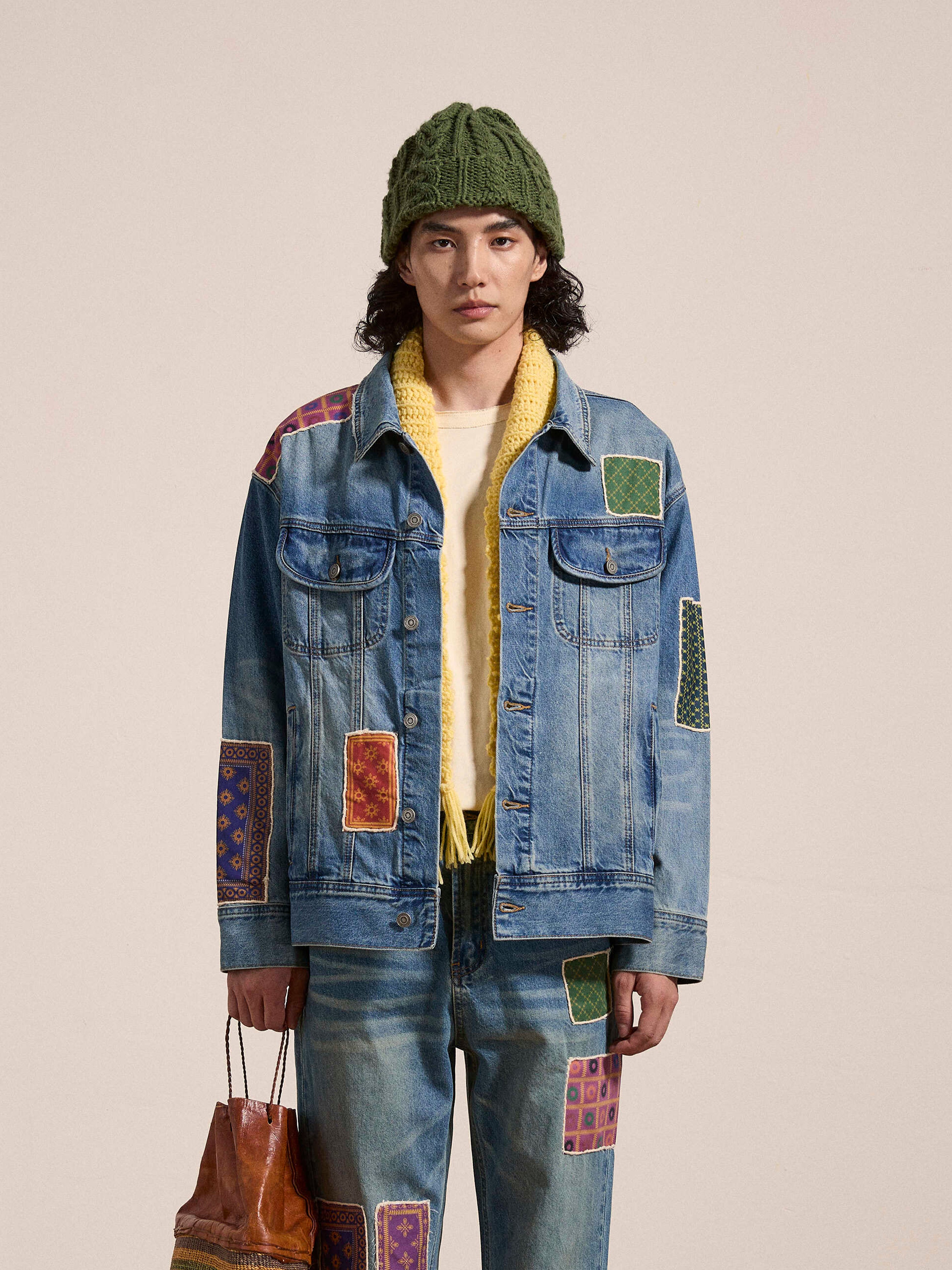 Person wearing a FOUND Kantha Patchwork Denim Trucker Jacket and jeans with South Asian inspired patches, a yellow sweater, and a green knit hat, holding a brown bag against a plain background.