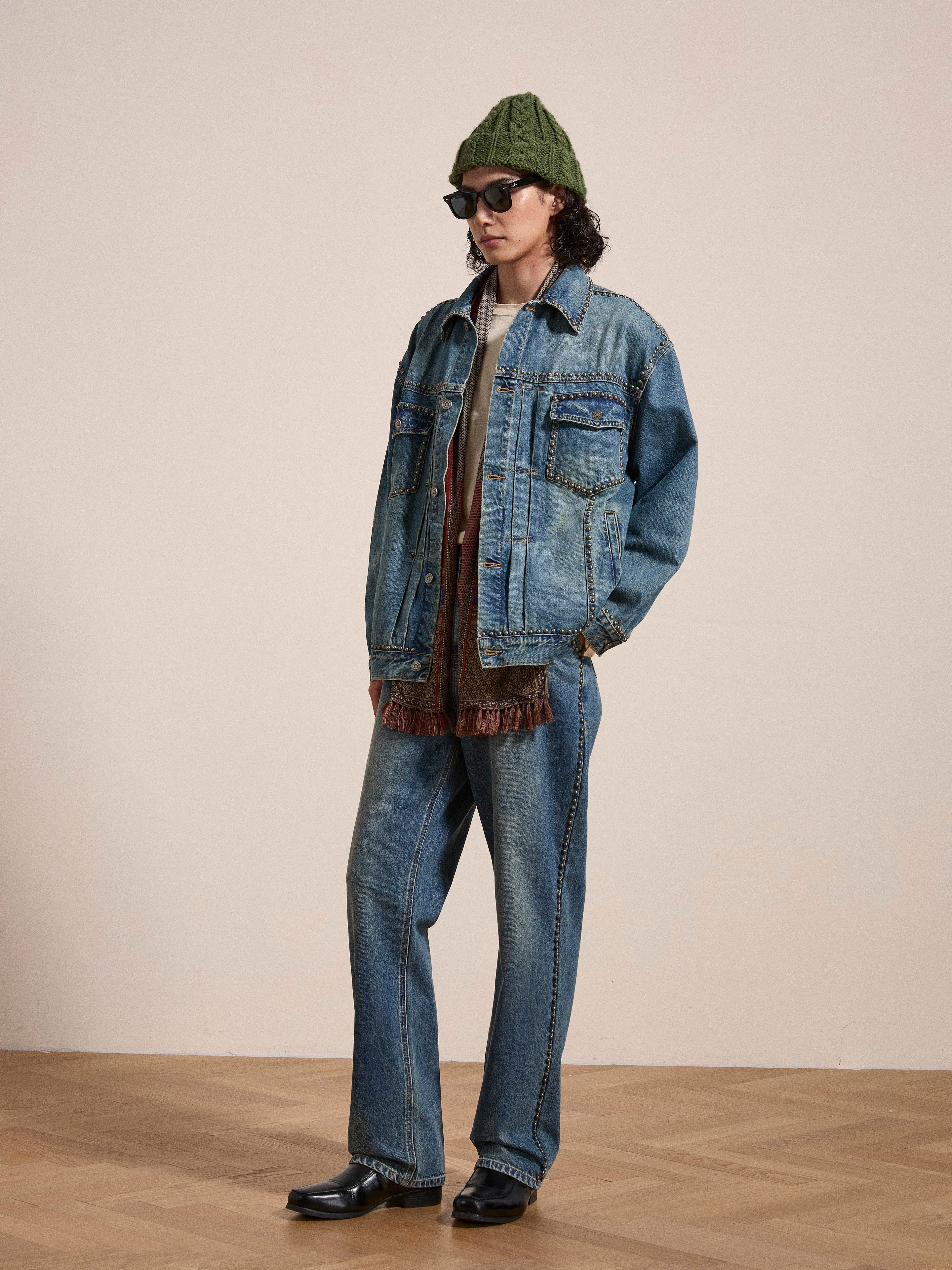 A person is standing indoors wearing a green knit cap, sunglasses, an Azar Studded Denim Trucker Jacket by FOUND in vintage blue wash with an oversized boxy fit, matching jeans, and black shoes. The background features a plain beige wall and wooden floor.