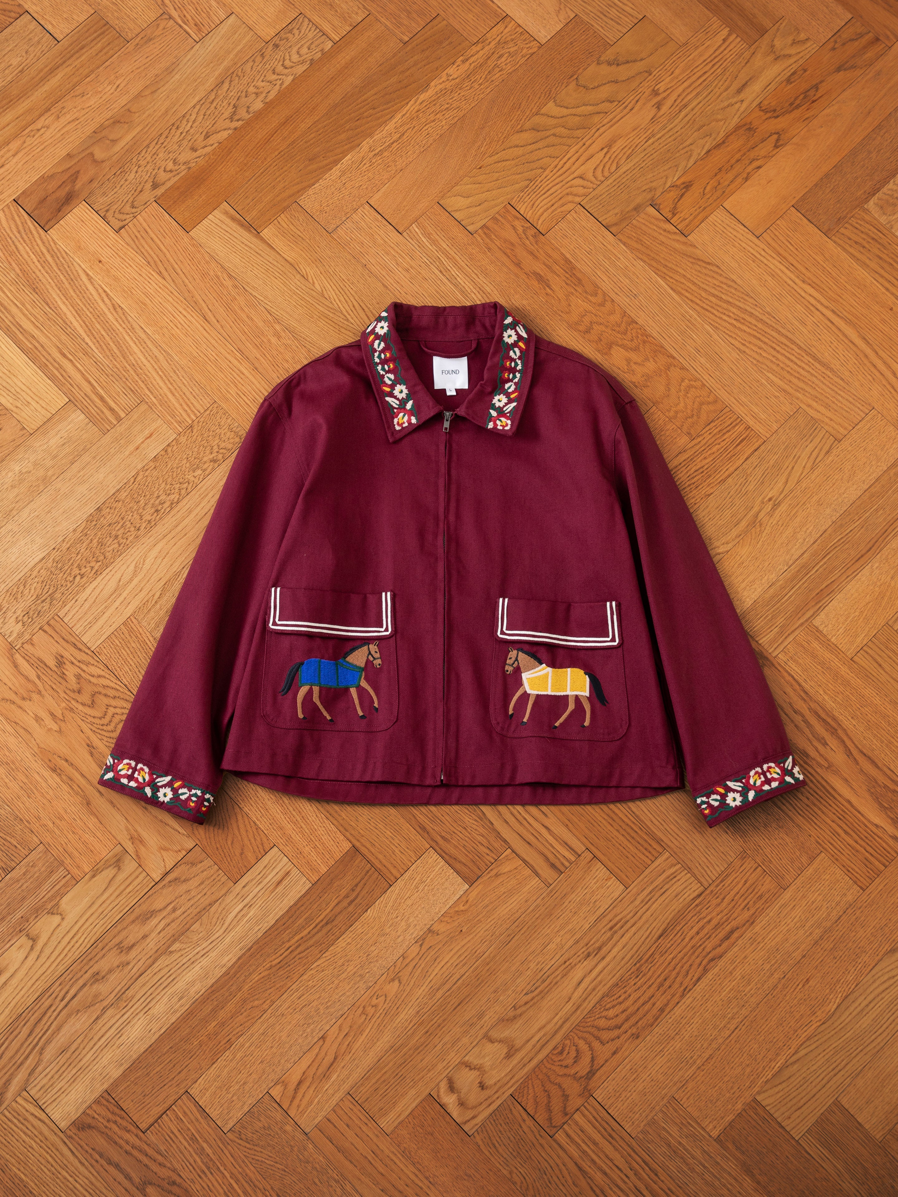 A maroon jacket with detailed embroidery, depicting horses and floral designs, laid flat on a wood parquet floor. This Oxblood Horse Equine Jacket by FOUND is reminiscent of 70's jockey uniforms, crafted from lightweight cotton twill for comfort and style.