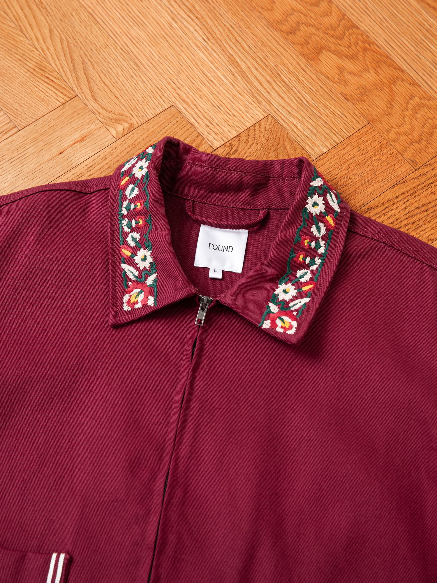 An Oxblood Horse Equine Jacket by FOUND, adorned with floral embroidery on the collar, lies on a wooden floor. Crafted from lightweight cotton twill, this piece captures the elegance of 70's jockey uniforms. Inside, a white tag simply reads 