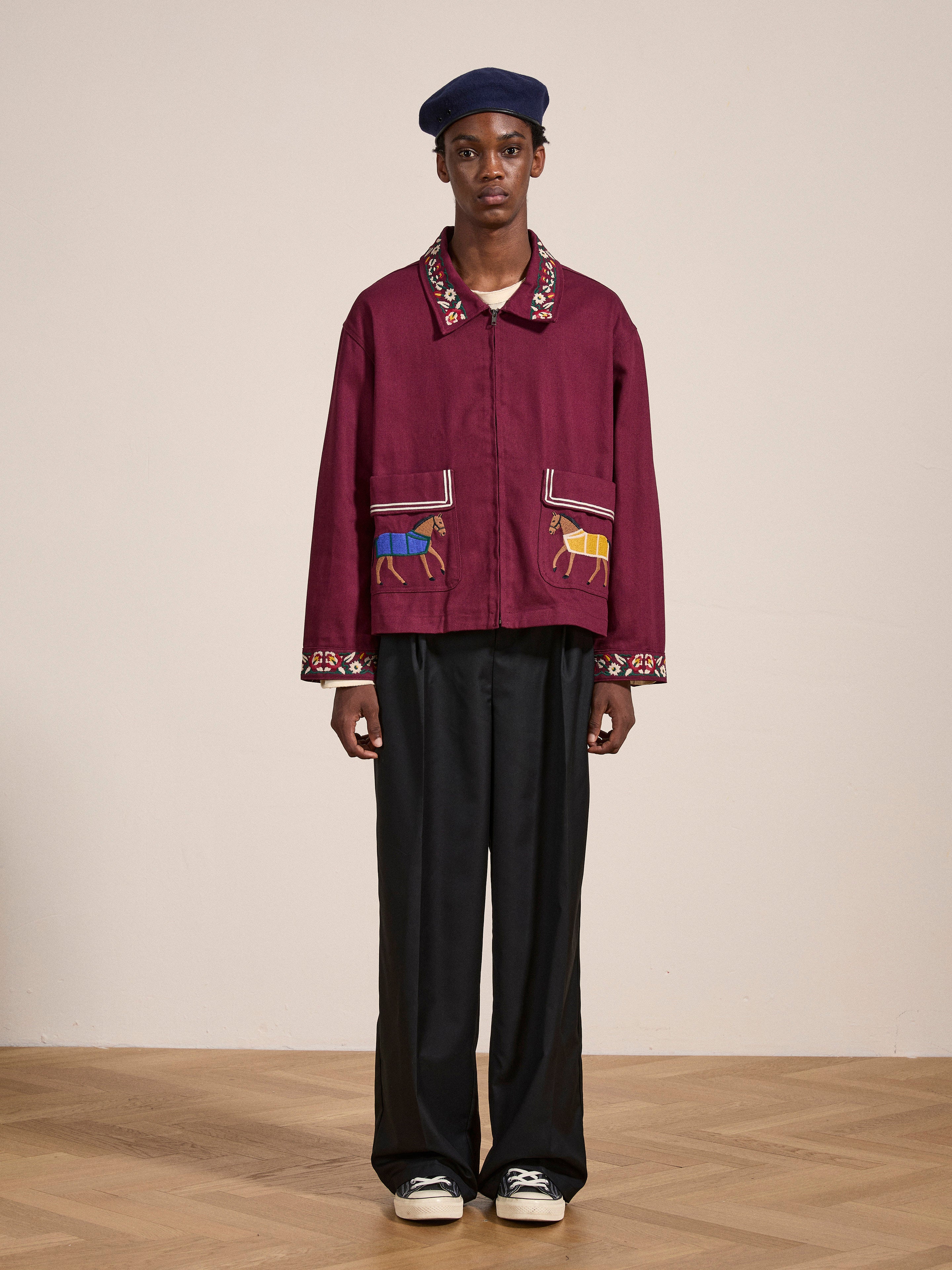 A person stands against a plain background, wearing an Oxblood Horse Equine Jacket by FOUND, featuring colorful embroidery on the collar and pockets. They complete the look with black wide-leg pants, a navy beret, and black sneakers.