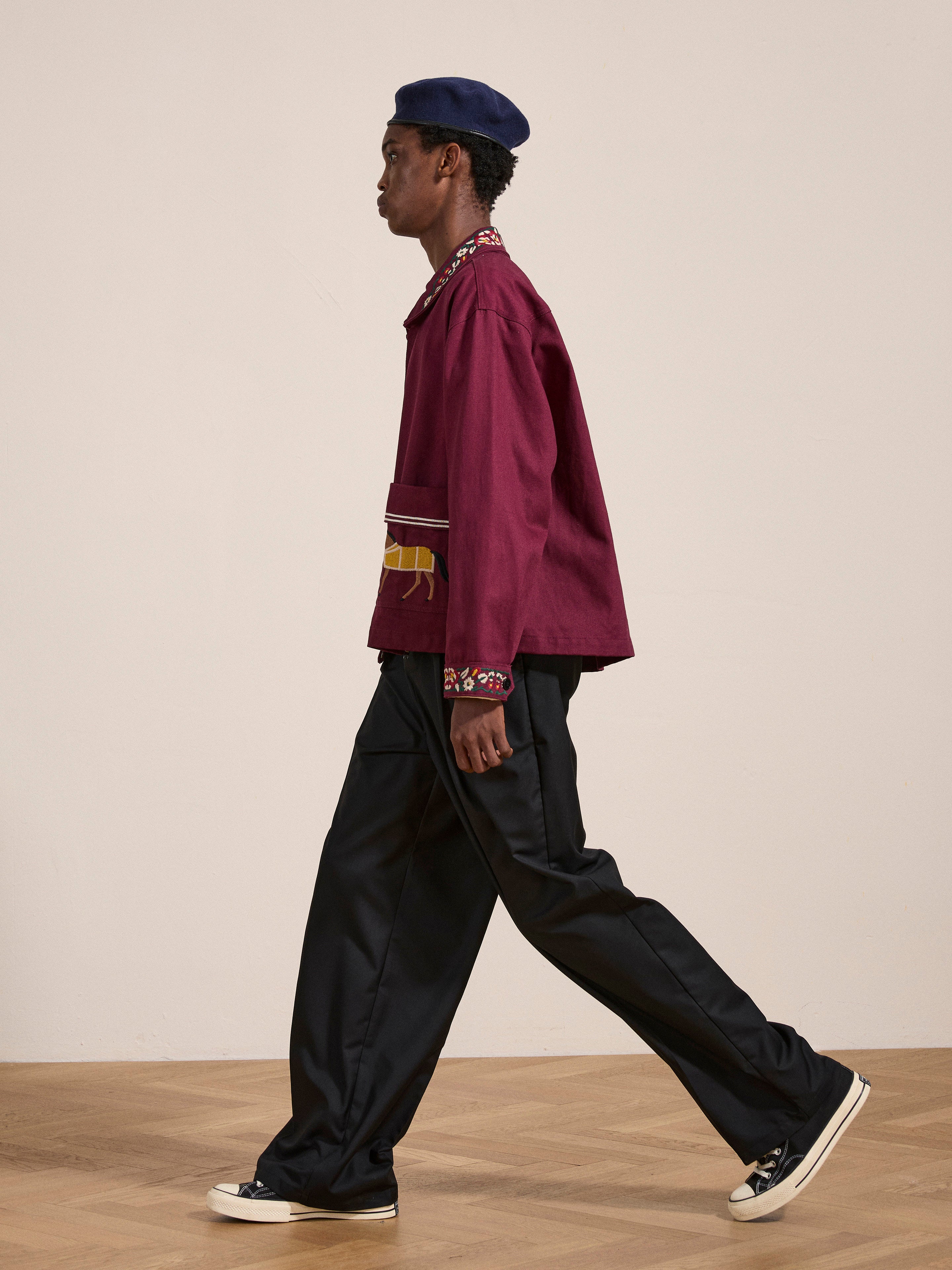 A person in an Oxblood Horse Equine Jacket by FOUND, featuring embroidered cuffs, paired with a black beret walks to the left, evoking the style of 70's jockey uniforms. They wear black pants and white sneakers. The background is plain with a wooden floor.