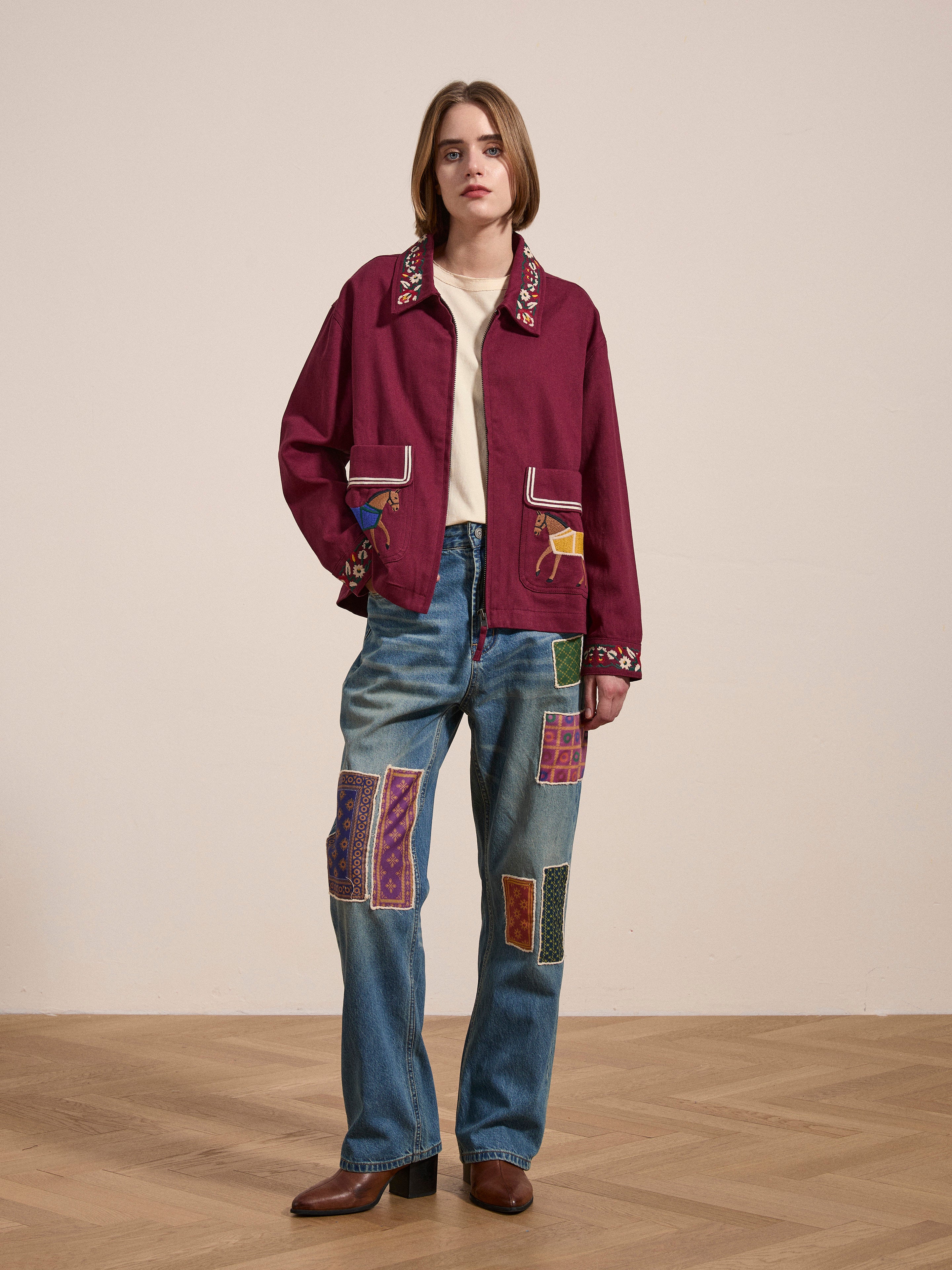 A person stands indoors on a wooden floor, dressed in a FOUND Oxblood Horse Equine Jacket with embroidery, complemented by a beige shirt, blue jeans featuring colorful patches inspired by 70's jockey uniforms, and brown shoes.