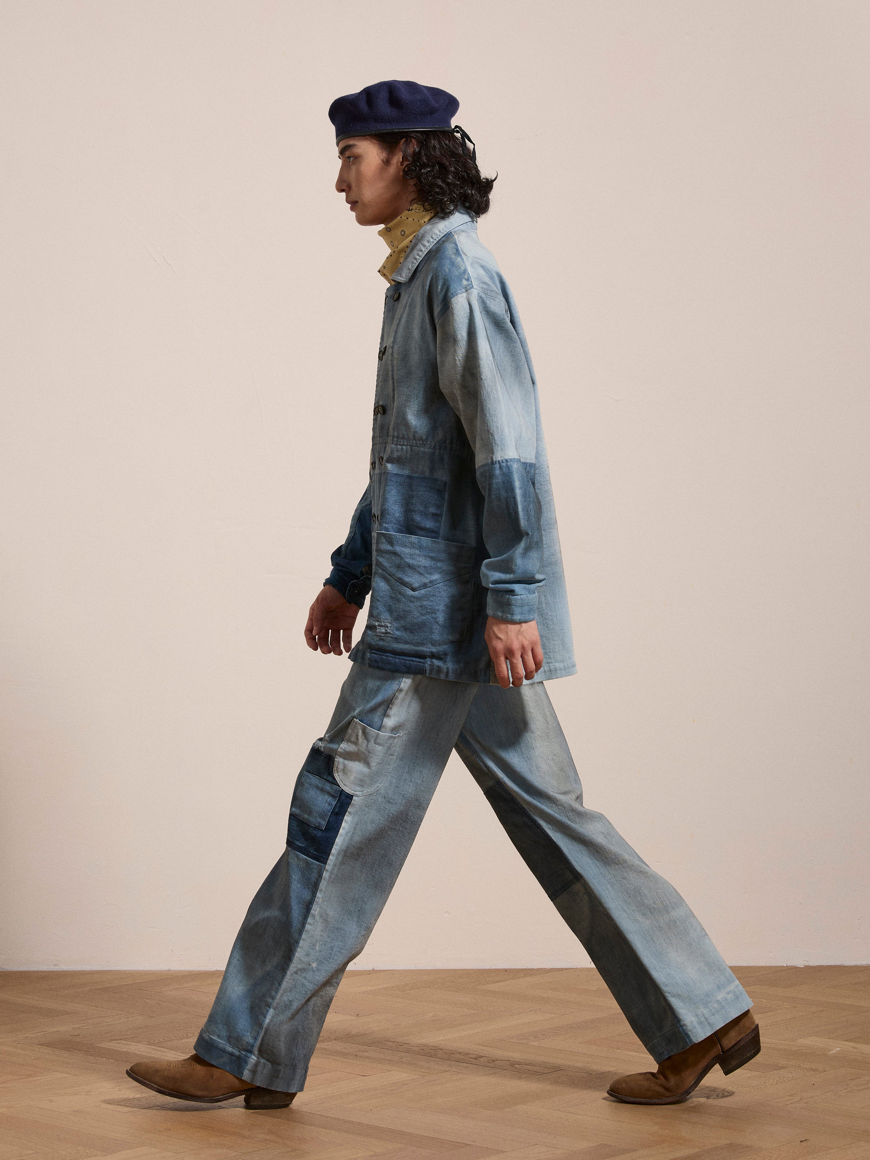 A person in Multi Patch Painters Work Pants by FOUND, reminiscent of antique French painter uniforms, and a dark beret walks across a wooden floor.