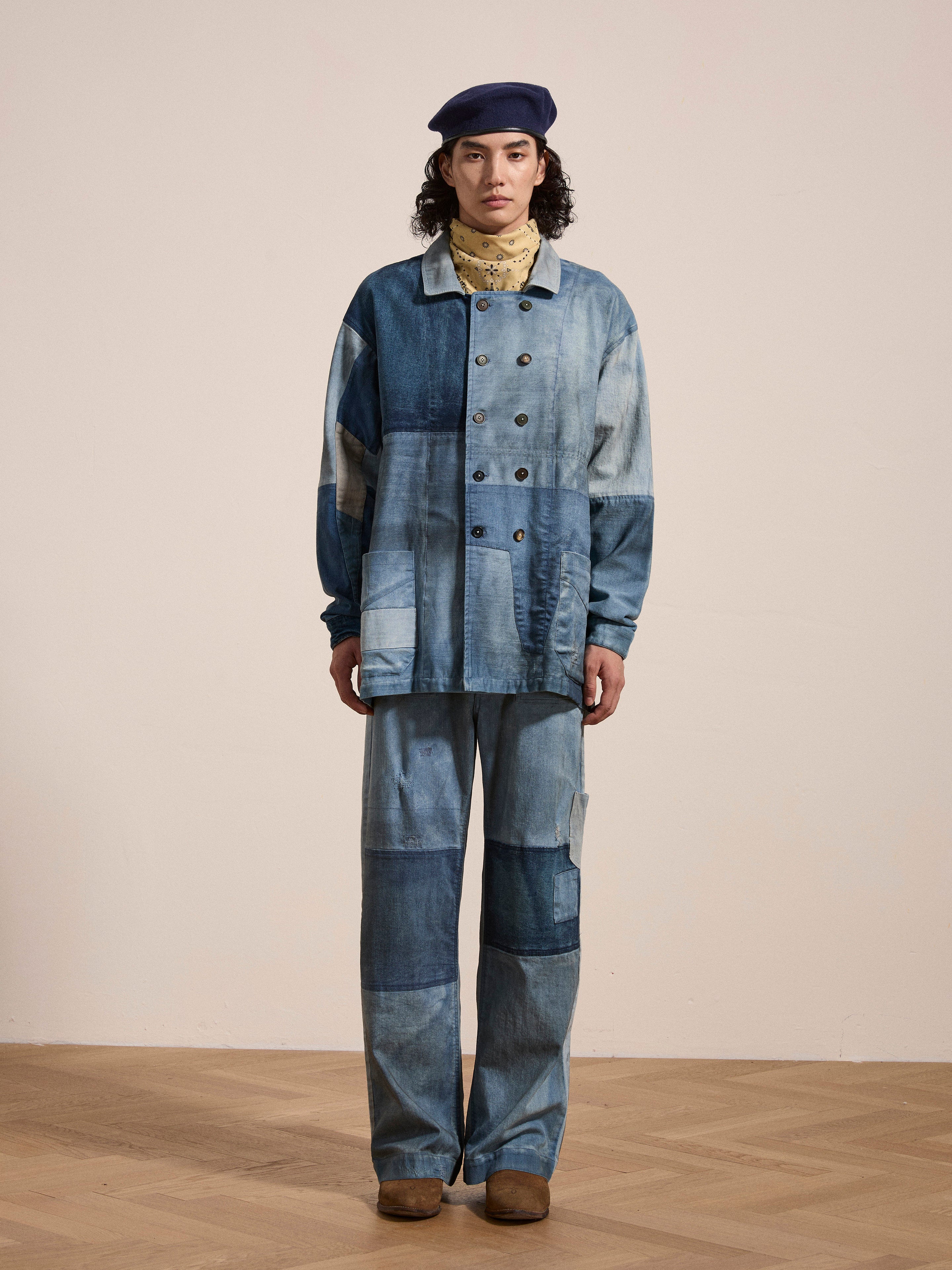 A person wearing the FOUND Multi Patch Painters Work Jacket paired with matching denim pants, a beige scarf, and a navy beret, standing on a light wood floor against a plain wall.