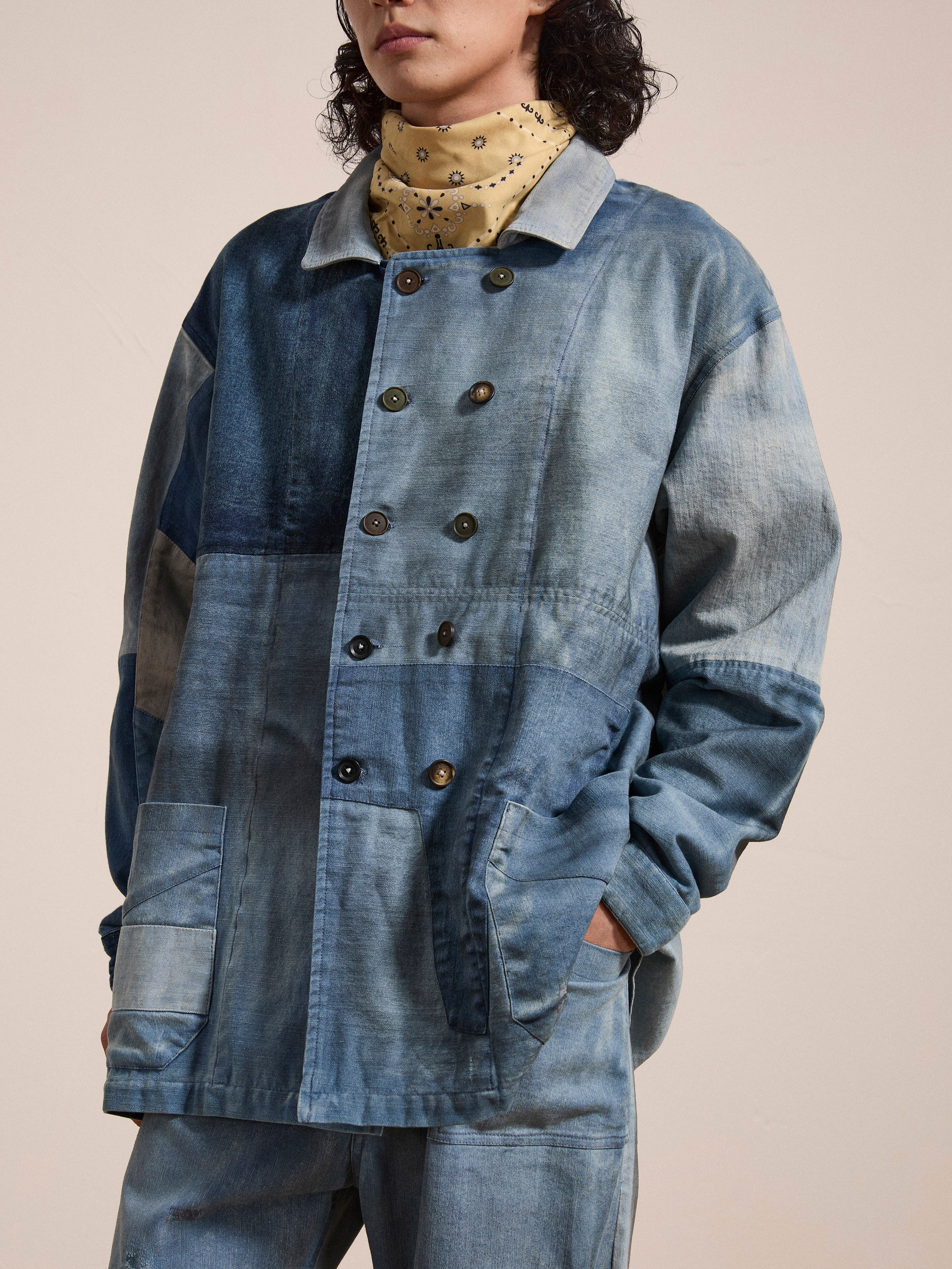 A person wearing the FOUND Multi Patch Painters Work Jacket, an oversized button-up denim piece with hand distressing in a blend of light and dark blue patches reminiscent of an antique French Painter's Work Jacket. The jacket sports several pockets, and the person also wears a patterned scarf around their neck.