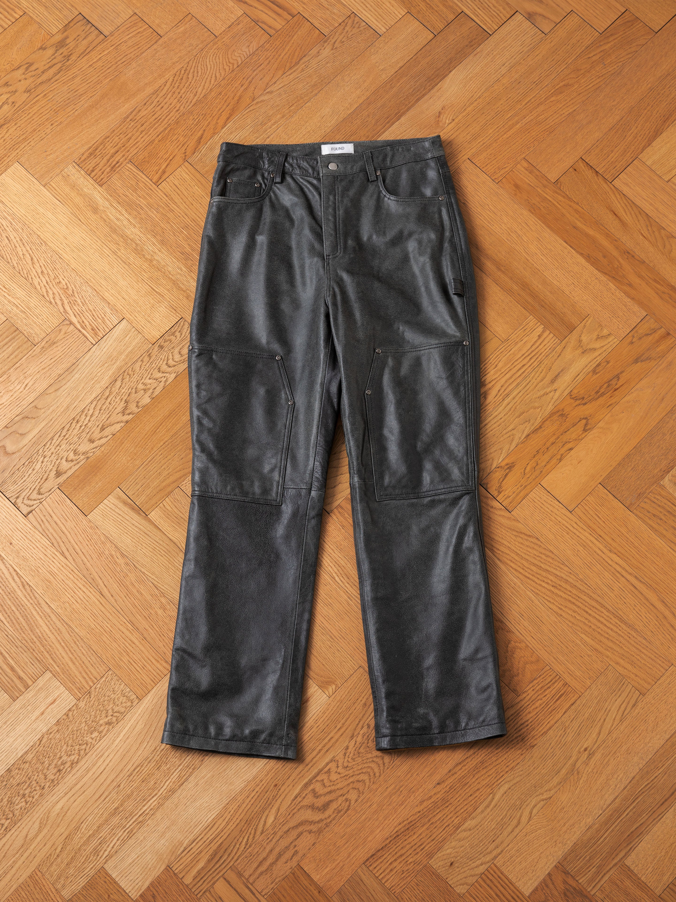 Distressed Genuine Leather Carpenter Pants by FOUND with multiple pockets lying flat on a parquet wooden floor.