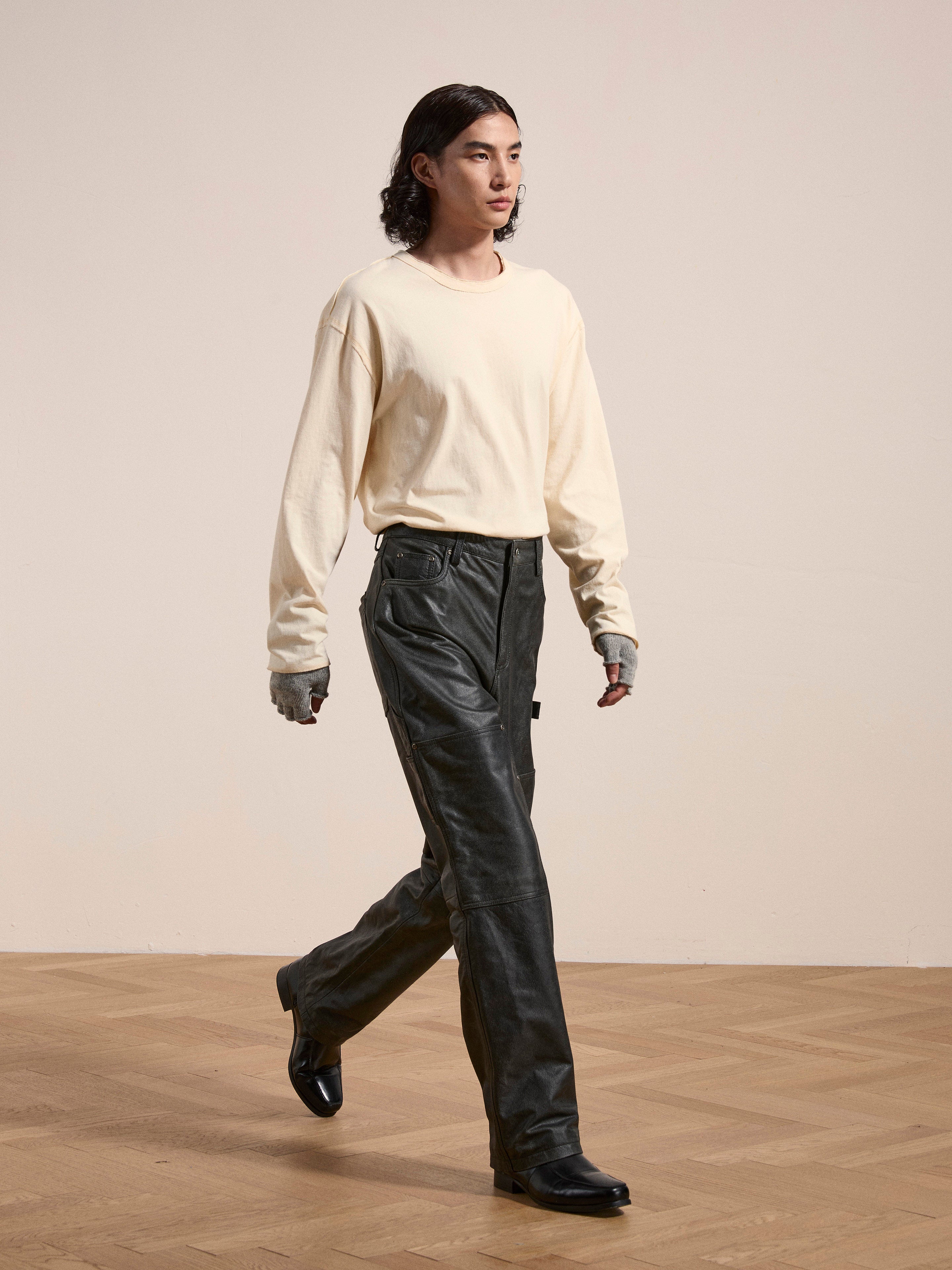 A person with long dark hair walks on a wooden floor, wearing a cream long-sleeve shirt, FOUND Distressed Genuine Leather Carpenter Pants in a double-knee silhouette, and black shoes.