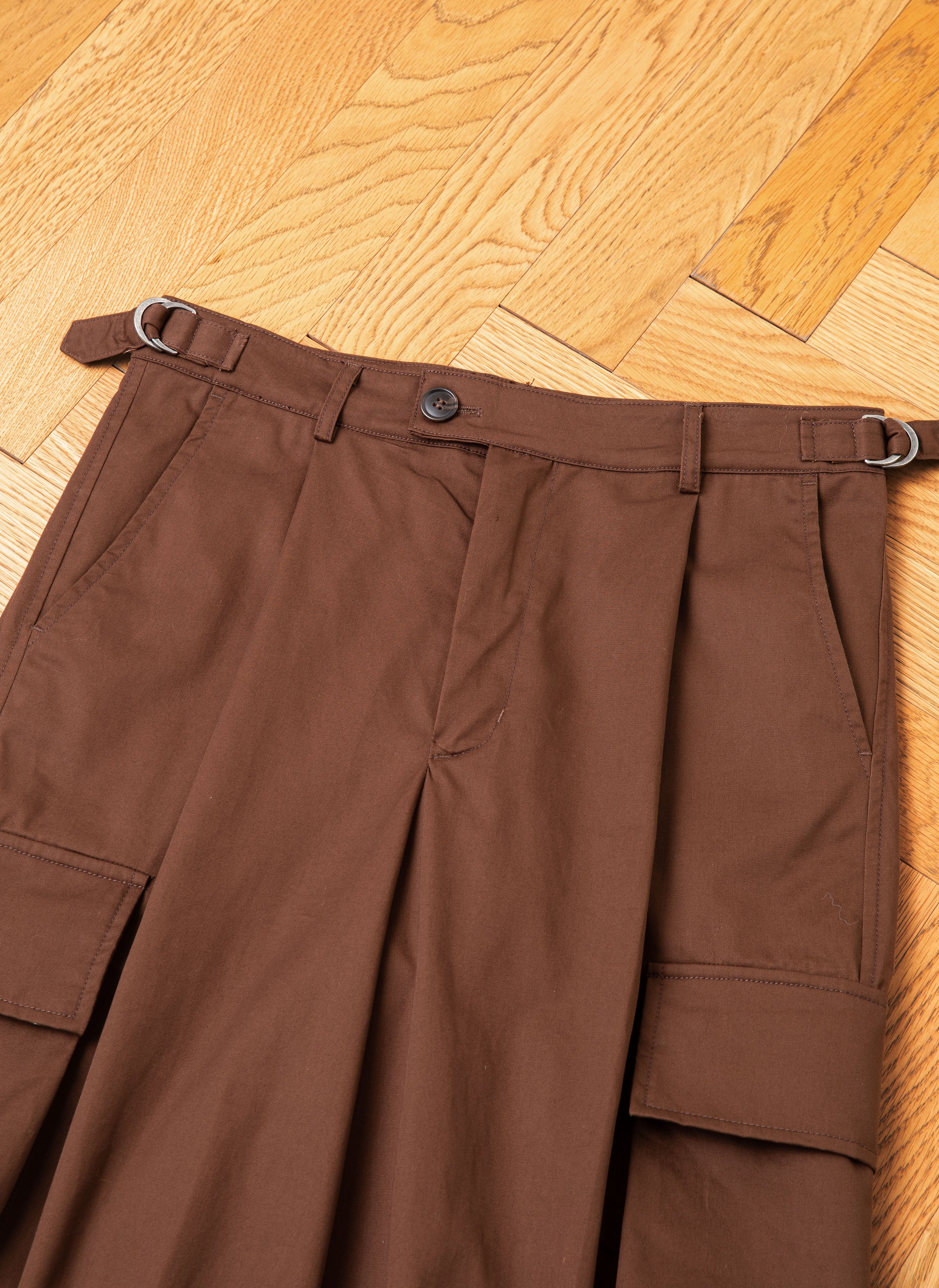 A pair of FOUND Pleated Pocket Trousers, featuring a tailored military style with side cargo pockets and belt loops, lies on a wooden floor.