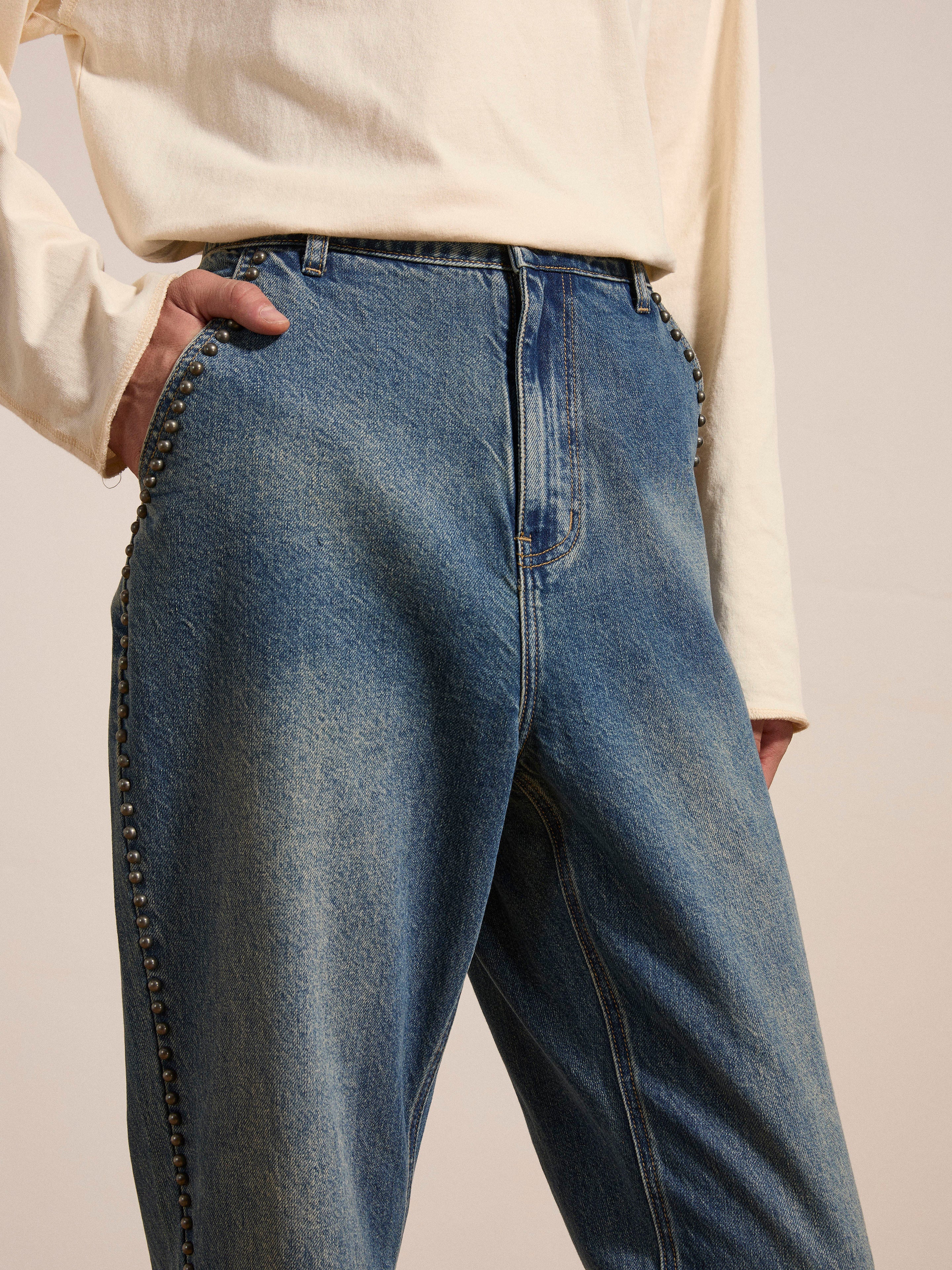 A person wearing FOUND's Azar Studded Jeans in a vintage blue wash, adorned with antique matte silver studs, and a long-sleeved beige shirt stands with one hand in their pocket.