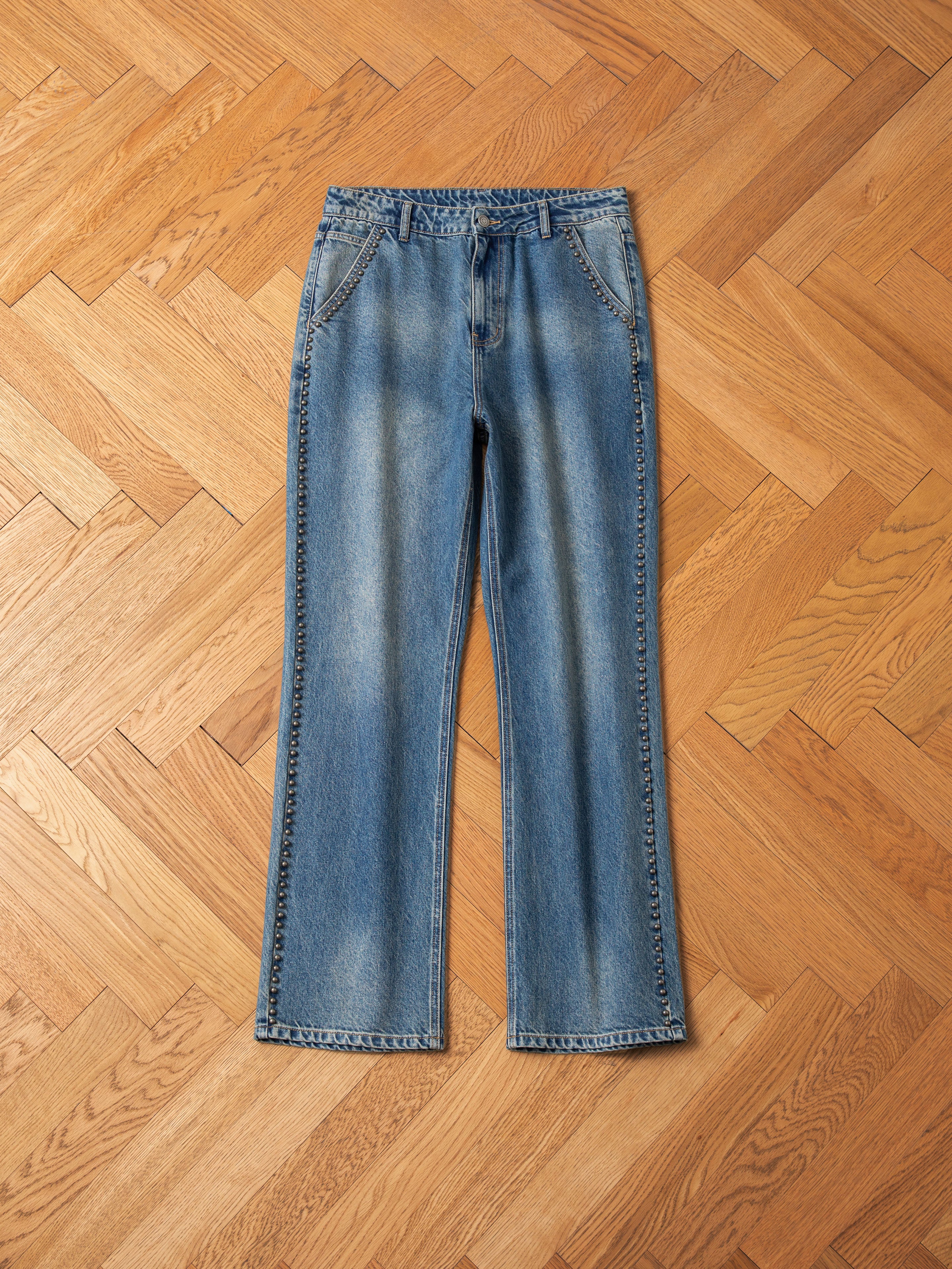 A pair of Azar Studded Jeans by FOUND in a vintage blue wash, featuring slightly faded detailing and antique matte silver studs, laid flat on a wooden herringbone-patterned floor.