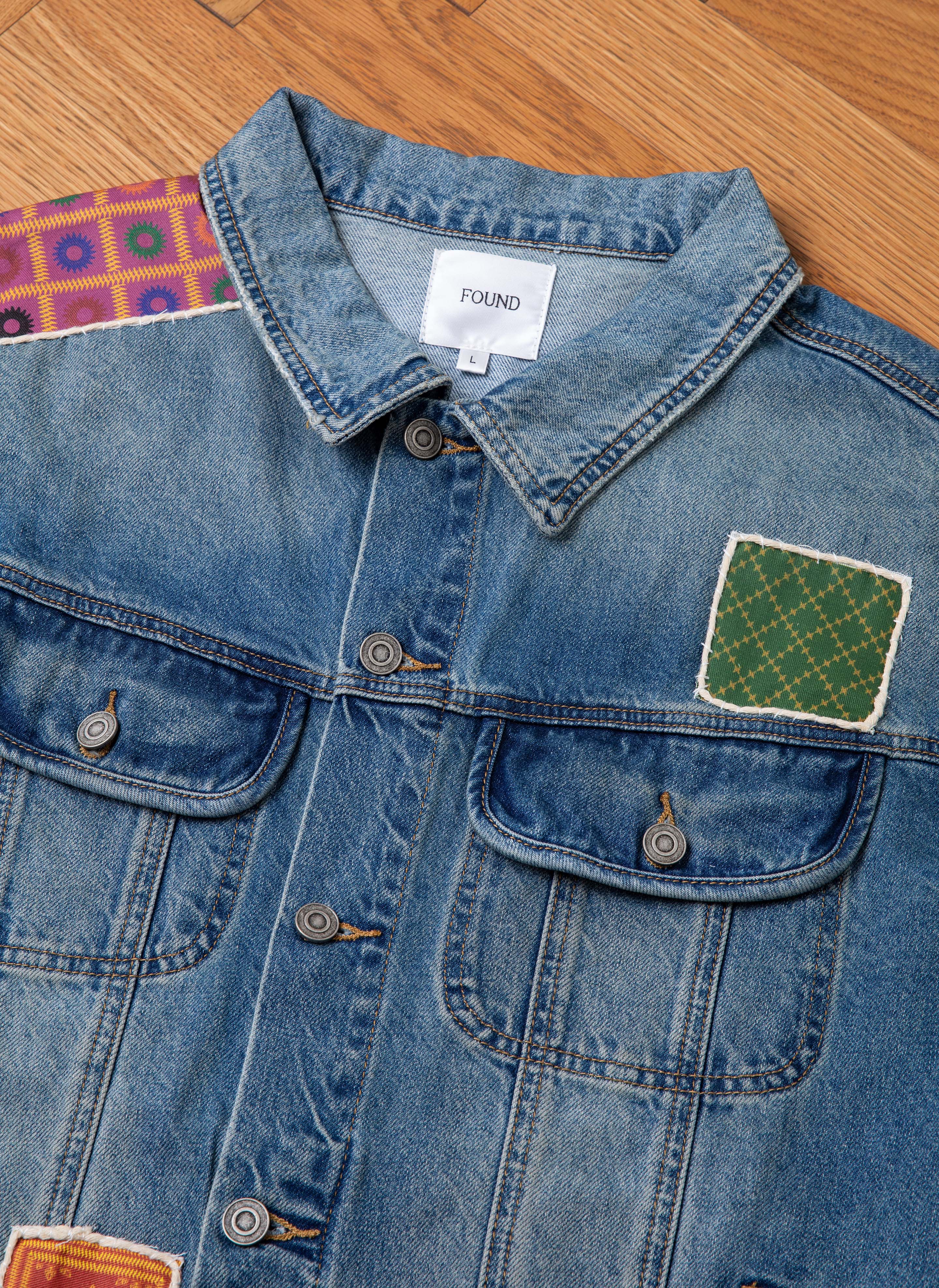 Close-up of a Kantha Patchwork Denim Trucker Jacket by FOUND. The jacket features buttoned chest pockets and is adorned with South Asian inspired patches on the shoulder and chest. The wooden floor is visible in the background.