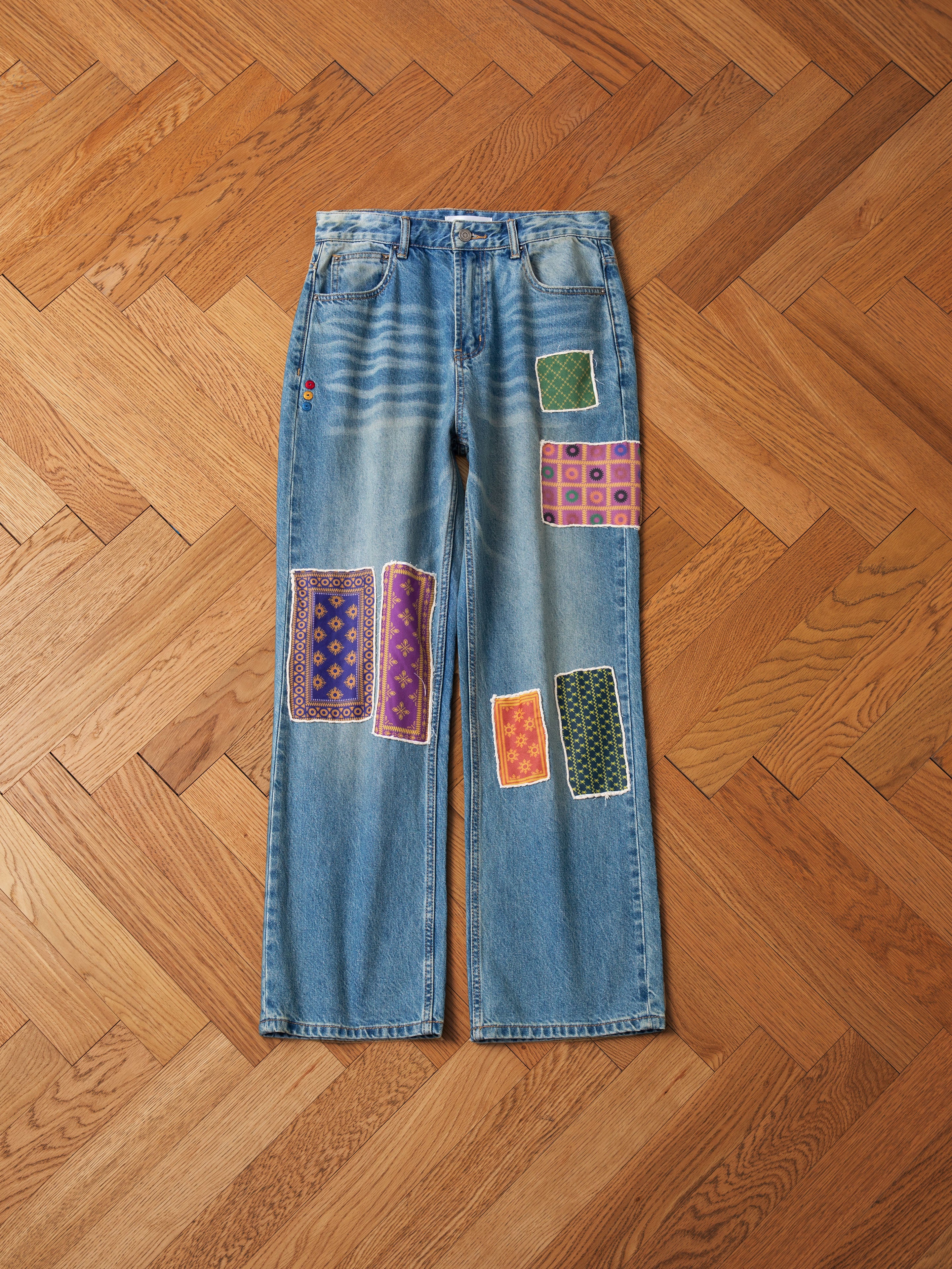 A pair of Found's Kantha Patchwork Jeans, featuring vibrant South Asian-inspired patches, beautifully showcased on a wooden herringbone floor.